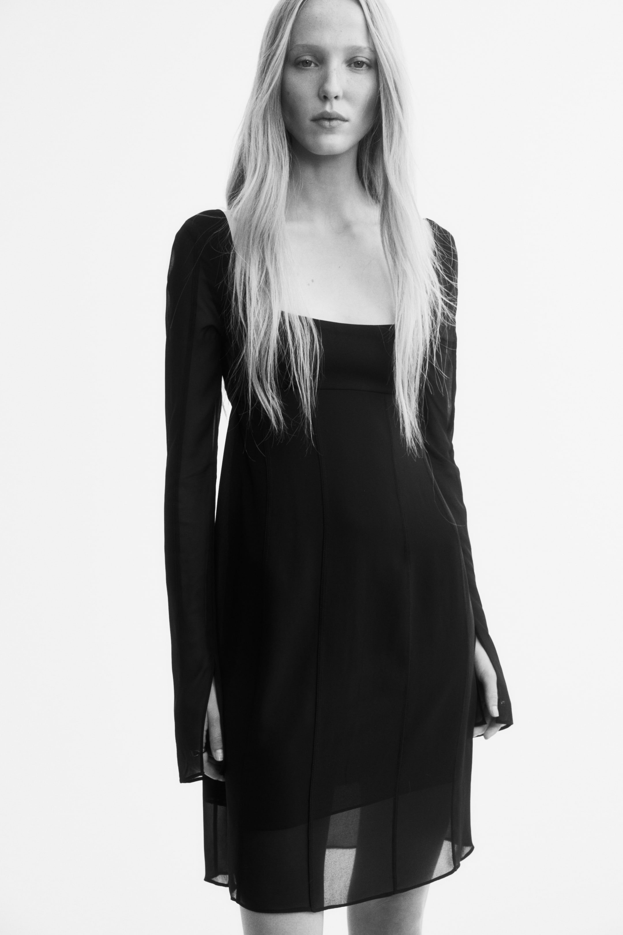 Größeres Bild anzeigen: Das Model trägt ein knielanges, figurbetontes schwarzes Kleid mit einem tiefen, quadratischen Ausschnitt und langen Ärmeln mit Schlitzmanschetten. Das Kleid ist mit einer horizontalen Naht unterhalb der Brust, vertikal verlaufenden Nähten an der Vorderseite und einem transparenten Chiffon-Saum gearbeitet.
