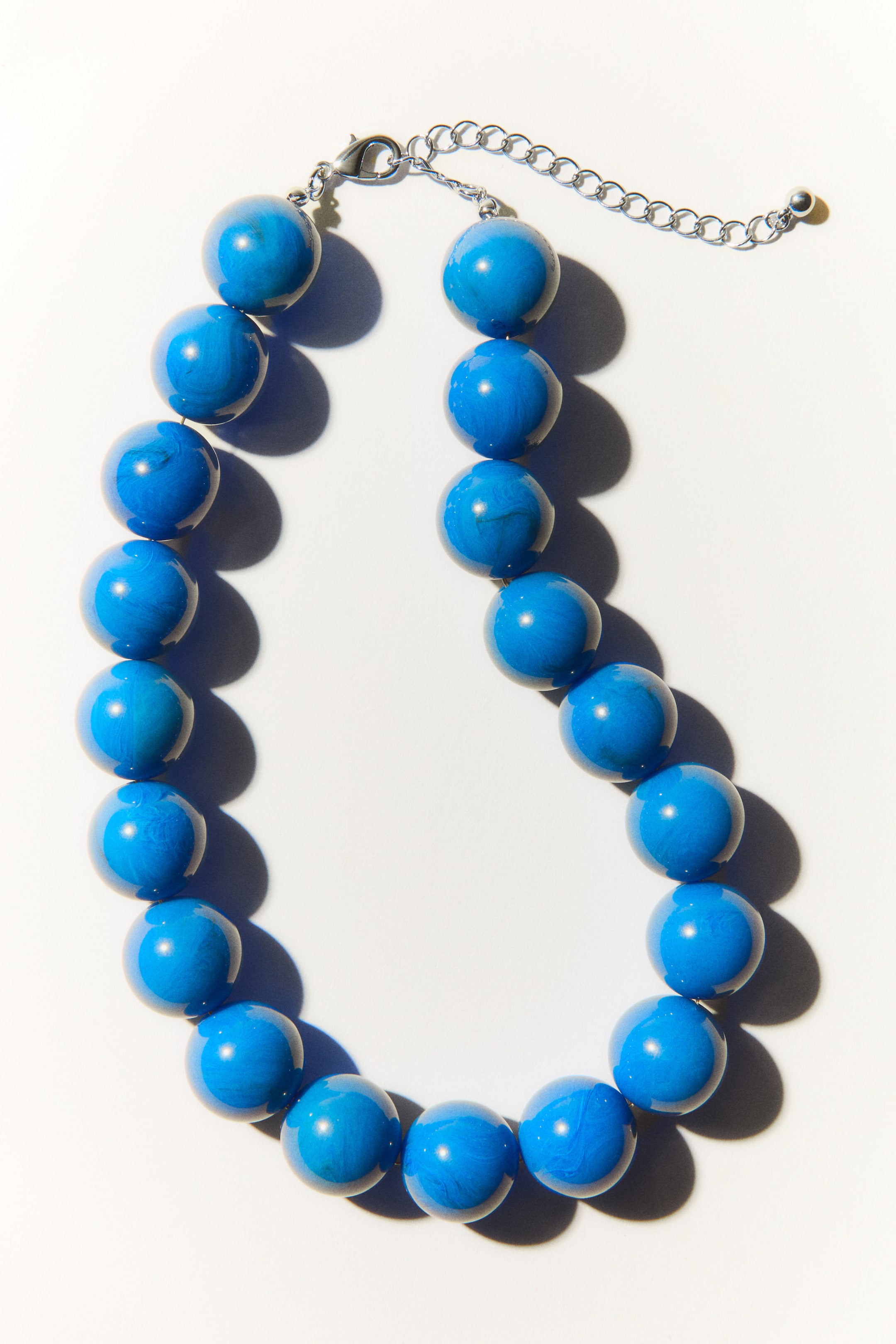 Ver imagen más grande: Un collar corto está ensartado con grandes cuentas de plástico azul, cada una mostrando un sutil patrón marmoleado. Una cadena de longitud ajustable se asegura con un cierre de gatillo de tono plateado.