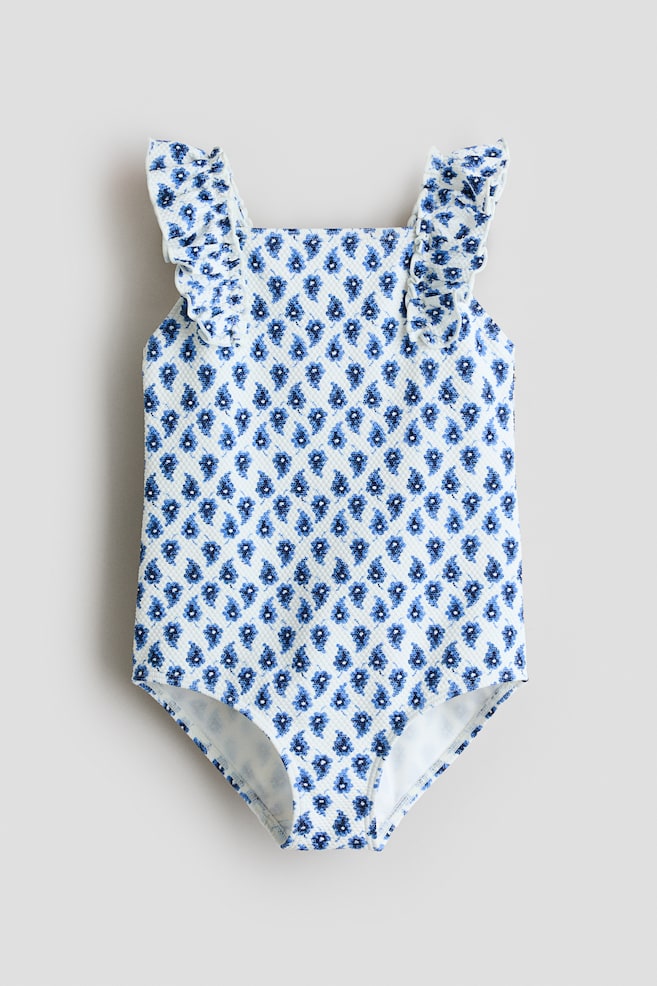 Un maillot de bain une pièce présente un tissu texturé blanc avec de petits motifs floraux bleus formant une grille géométrique. Il se distingue par des bretelles à volants, un décolleté carré, et est présenté de face. - 1