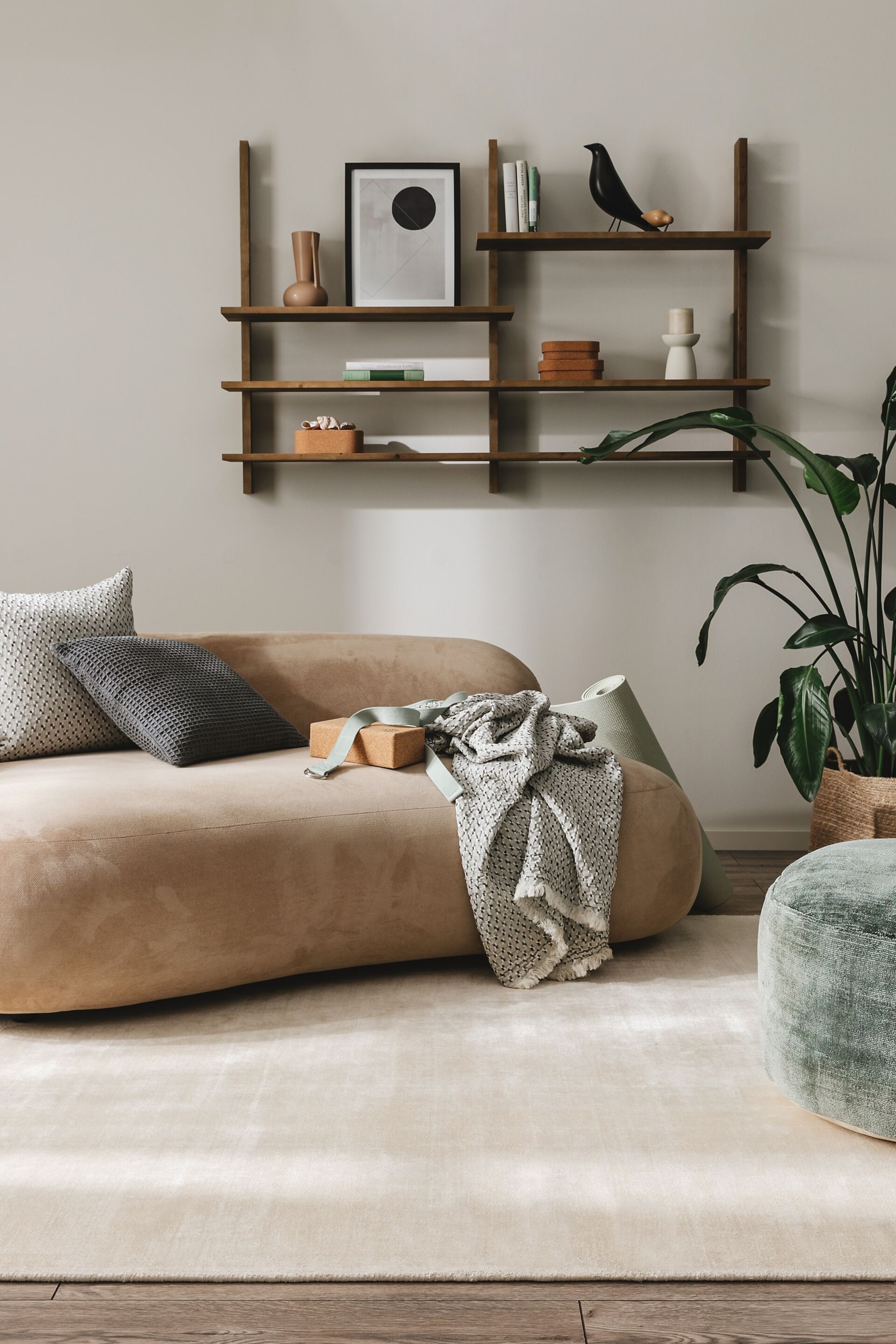 Größeres Bild ansehen: Das hellbraune Polstersofa zeichnet sich durch eine geschwungene, organische Form aus und ist leicht nach rechts geneigt. Zwei dekorative Kissen, eine strukturierte Decke, ein Kork-Yogablock und ein hellgrüner Yoga-Gurt liegen auf seiner Oberfläche, während eine passende Yogamatte an seiner Seite lehnt.