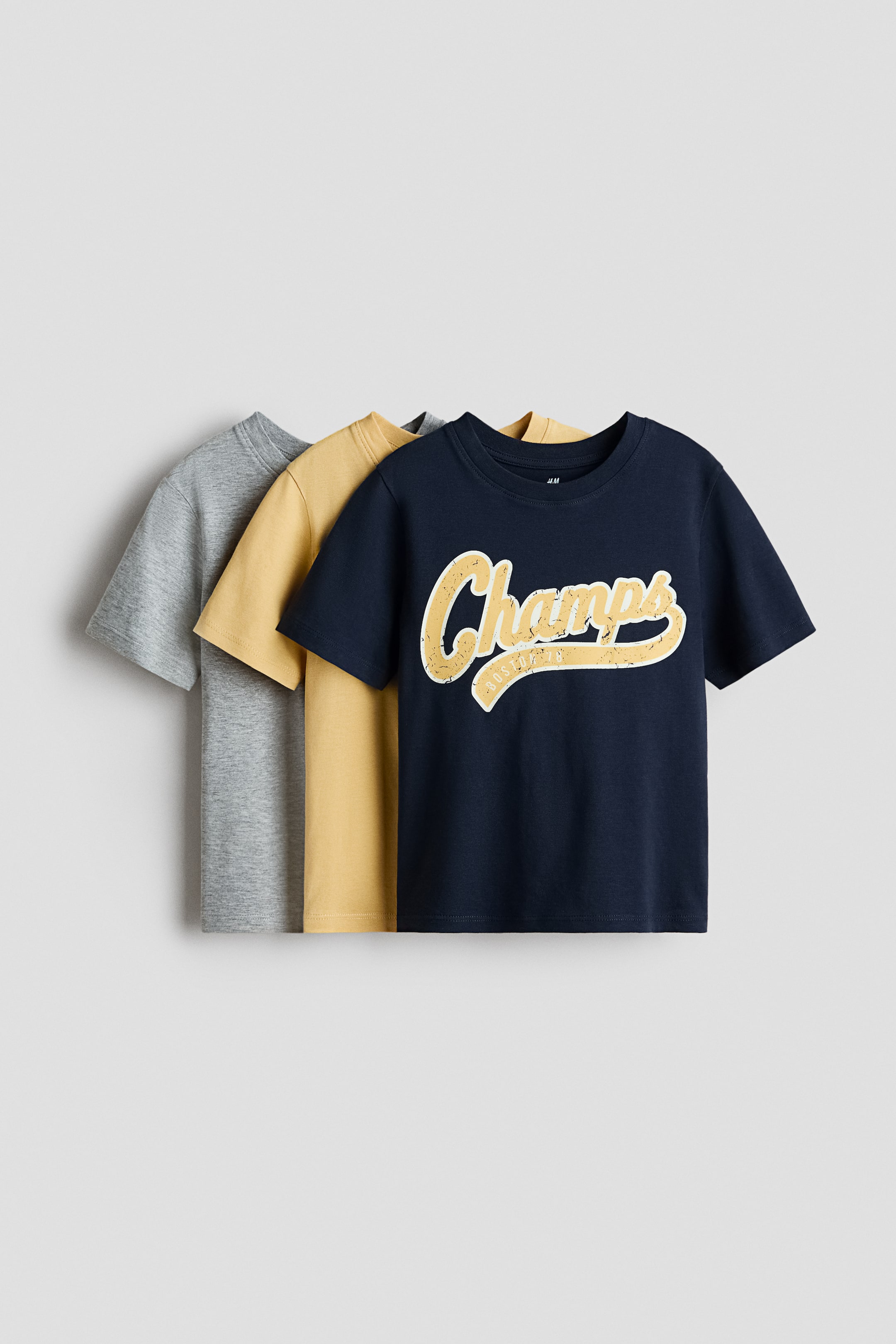 View larger image: Three short-sleeve t-shirts are layered, with the navy blue t-shirt in front, a mustard yellow one behind it, and a heather gray one at the back. The navy blue t-shirt features a distressed cream-colored "Champs" graphic with a thin white outline and "80s ATHLETIC" text on its front.