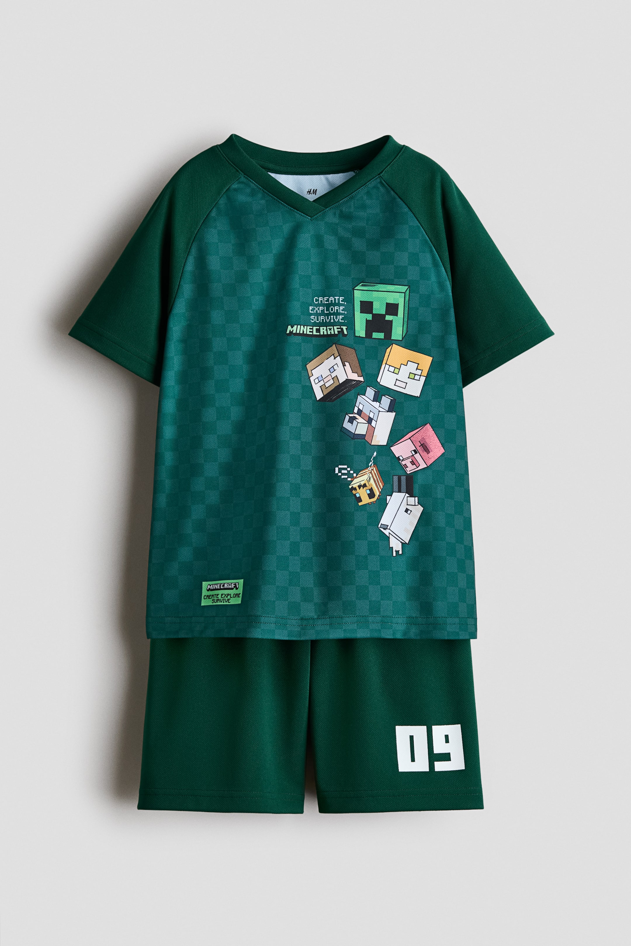 Grösseres Bild ansehen: Ein dunkelgrünes Fussball-Set wird von vorne gezeigt, mit einem Oberteil, das ein dezentes Karomuster auf dem Körper aufweist, einfarbige dunkelgrüne Raglanärmel, einen V-Ausschnitt und einen Aufdruck mit dem Text "CREATE, EXPLORE, SURVIVE. MINECRAFT" sowie verschiedenen Minecraft-Charakterköpfen. Die dazu passenden dunkelgrünen Shorts zeigen die Zahl "09" in Weiss auf dem linken Bein.