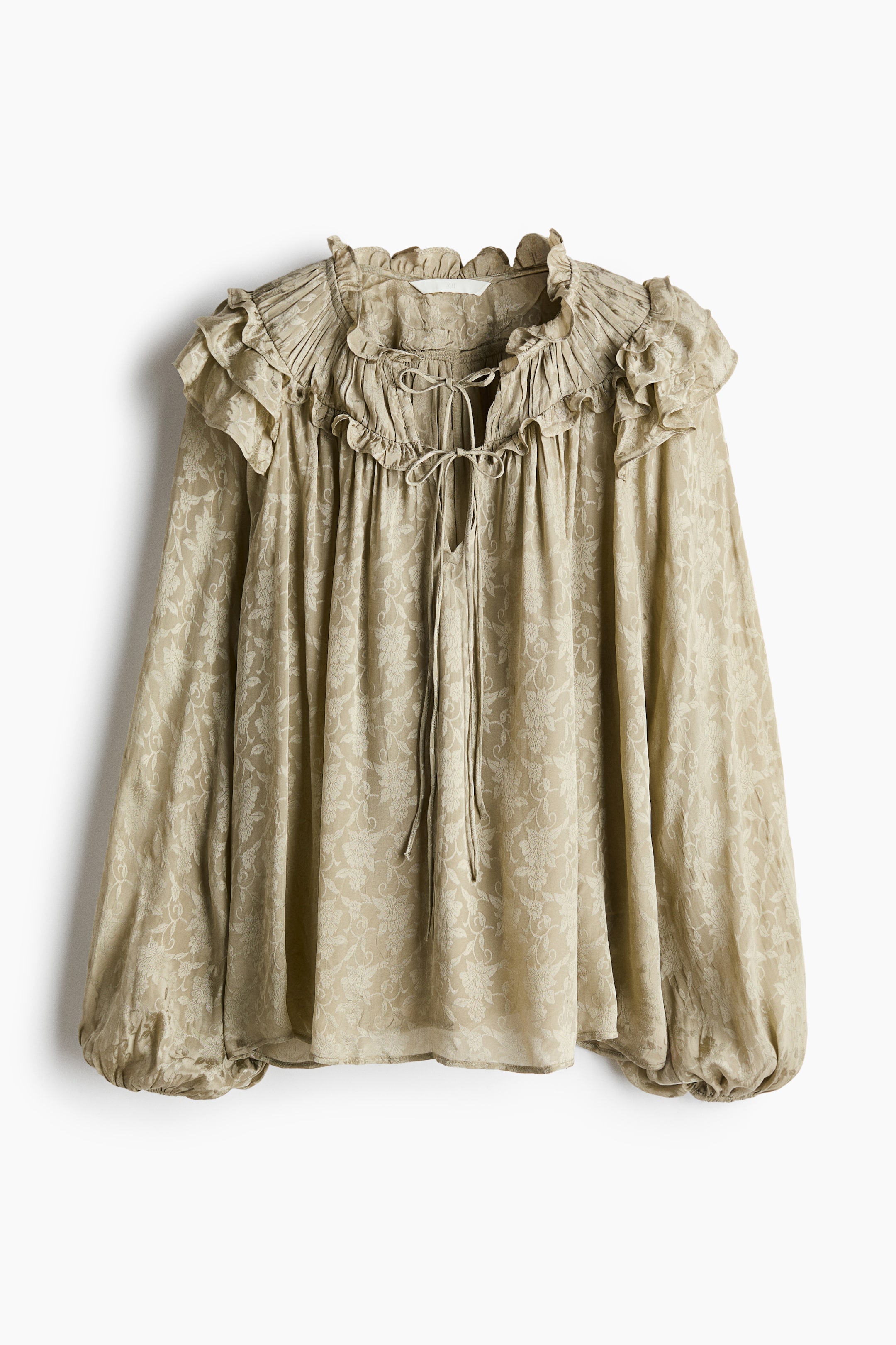 View larger image: A light beige blouse is shown from the front, featuring an all-over floral embossed pattern. It is designed with a high ruffled neckline, cinched with tie strings at the V-neck opening, and features long voluminous sleeves finished with gathered cuffs.