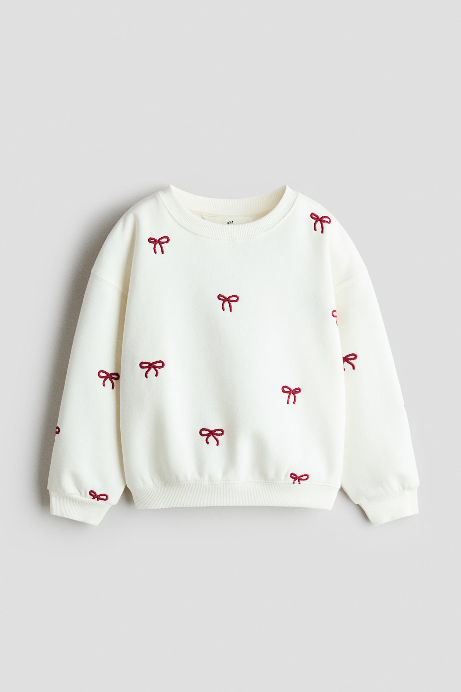 Krem renkli bir sweatshirt önden görülüyor; bisiklet yaka, düÅük omuzlar ve ribana manÅetler ile etek ucu detaylarına sahip. Sweatshirtün gövde ve kolları boyunca serpiÅtirilmiÅ küçük, nakıÅlı kırmızı fiyonk desenleri yer alıyor.