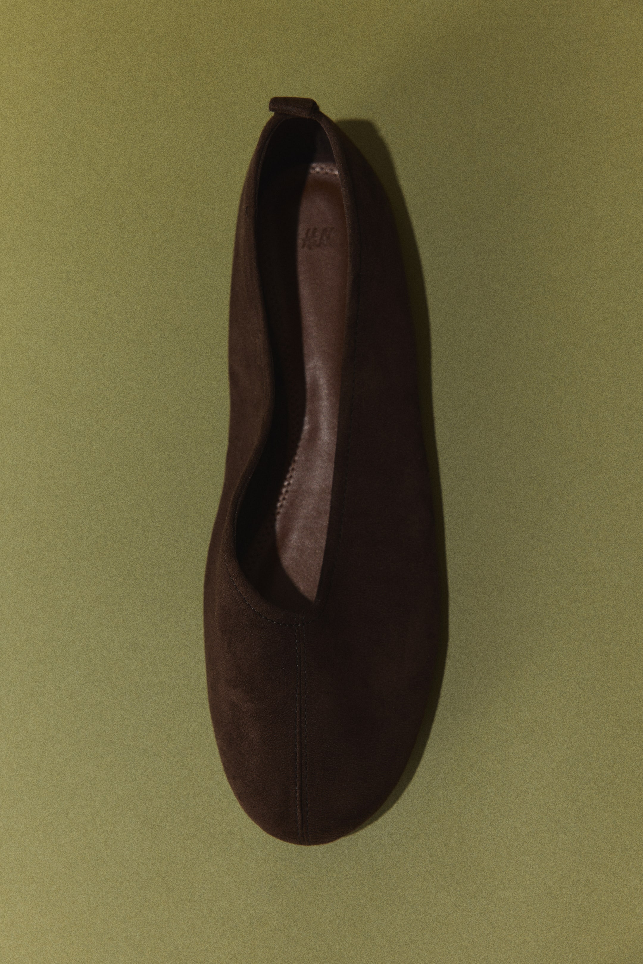 Größeres Bild ansehen: Ein dunkelbrauner flacher Schuh zeigt eine mandelförmige Spitze und ein V-förmiges Vorderblatt, gesehen aus der Draufsicht. Er verfügt über eine wildlederähnliche Oberfläche, eine kleine Zuglasche an der Ferse und eine sichtbare braune Innensohle mit eingeprägtem Logo.