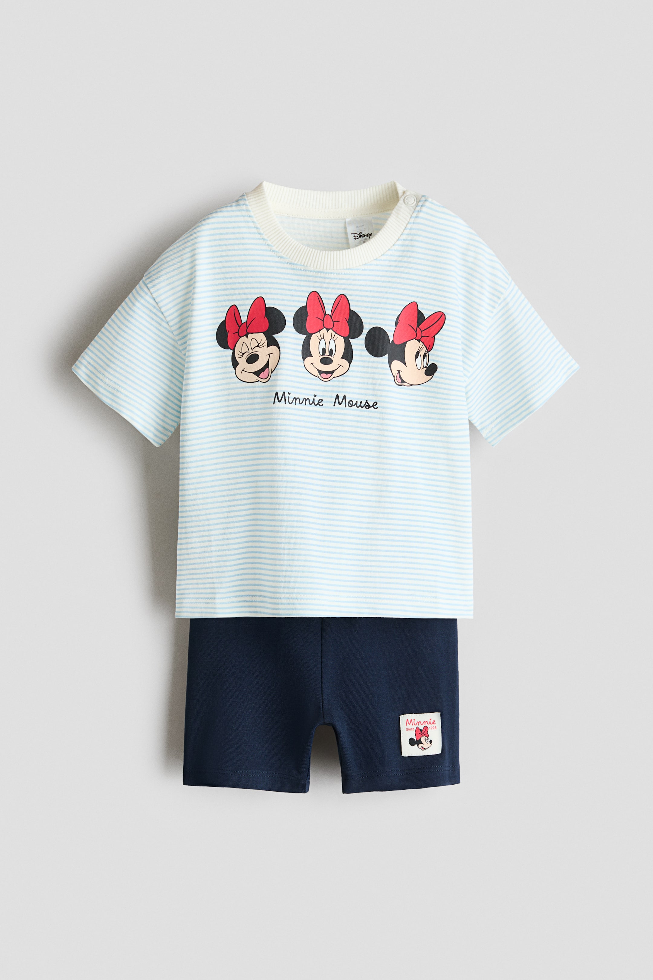 Größeres Bild anzeigen: Ein 2-teiliges Set aus bedruckter Baumwolle präsentiert ein hellblau-weiß gestreiftes Kurzarm-Oberteil mit geripptem Rundhalsausschnitt, Schulterdruckknöpfen und drei Minnie-Maus-Gesichtern mit dem Text „Minnie Mouse“ auf der Brust. Dunkelblaue Shorts vervollständigen das Set und verfügen über ein kleines Minnie-Mouse-Label am linken Bein vorne.