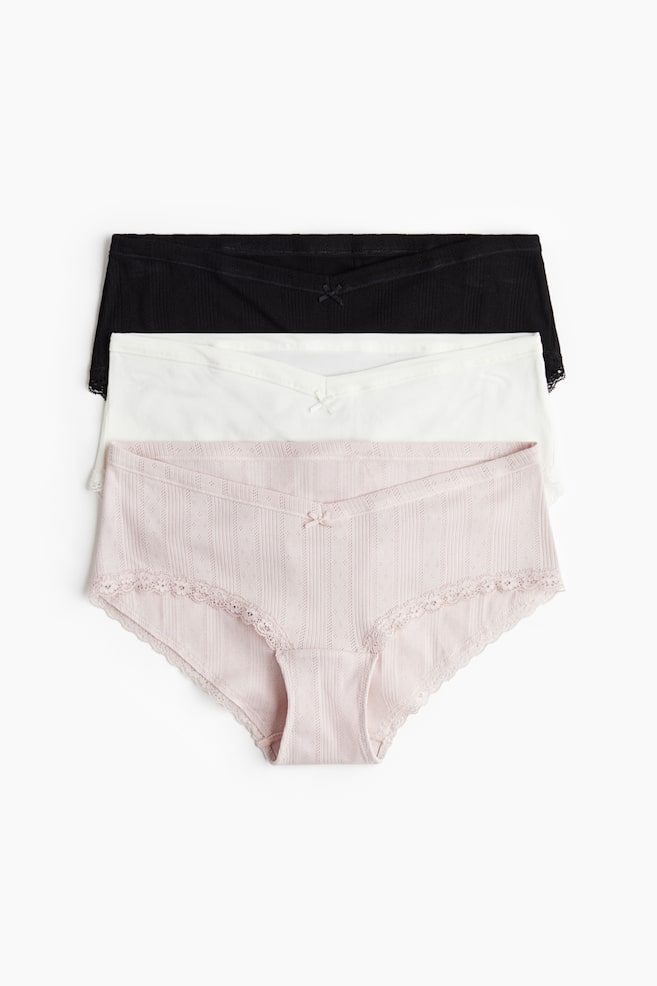 A stack of three low-rise briefs is presenting with their fronts visible. Each pair is featuring a V-shaped front waistband with a small bow and lace trim along the leg openings. The bottom light pink brief is displaying a textured fabric with vertical stripes and small eyelets, while the white and black briefs above it are showing ribbed textures. - 1