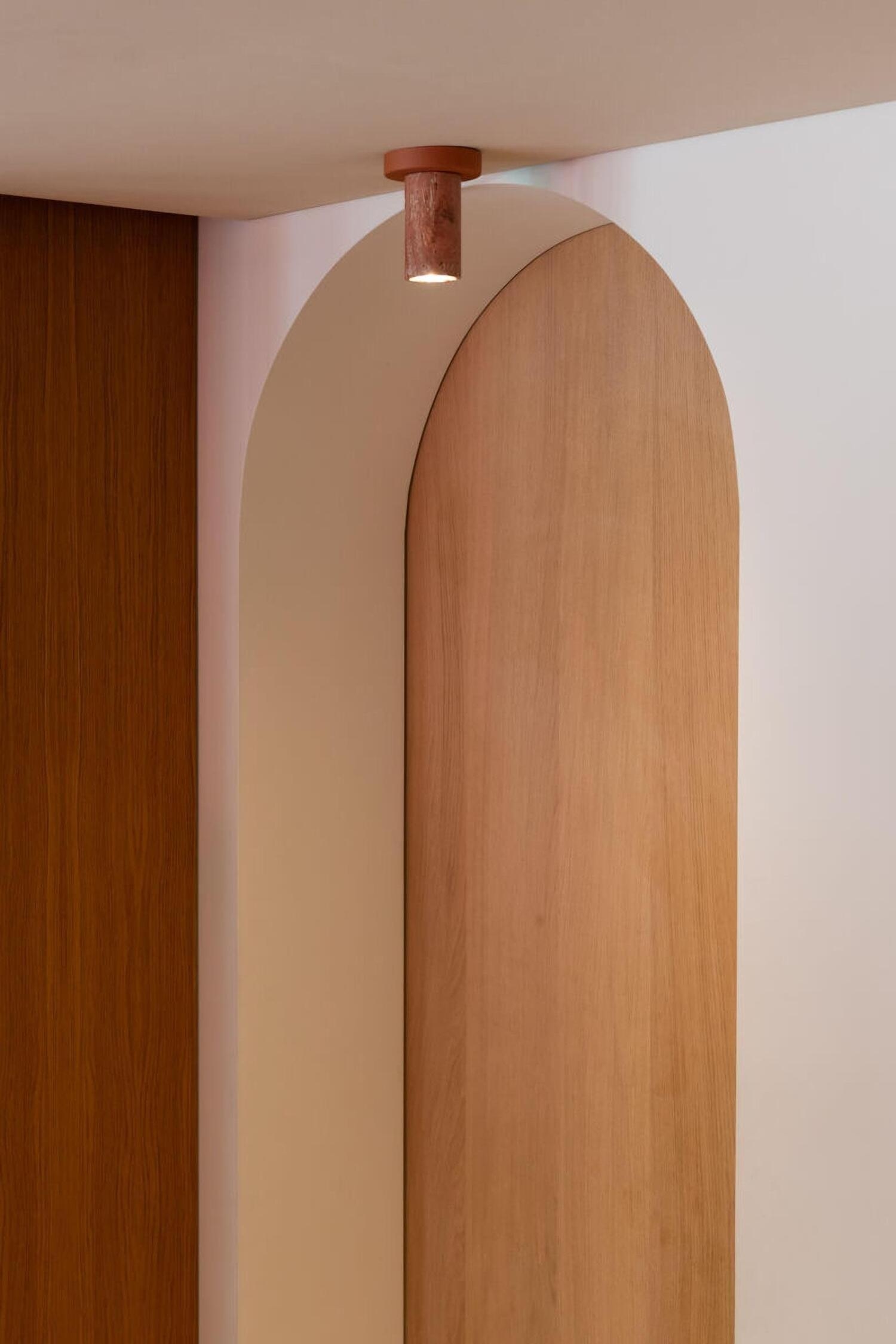Een aan het plafond gemonteerde verlichtingsarmatuur met een gladde, ronde terracotta afdekplaat en een cilindrische behuizing vervaardigd uit getextureerde, roodbruine travertijnsteen. Uit de onderste opening van de stenen cilinder straalt warm licht.