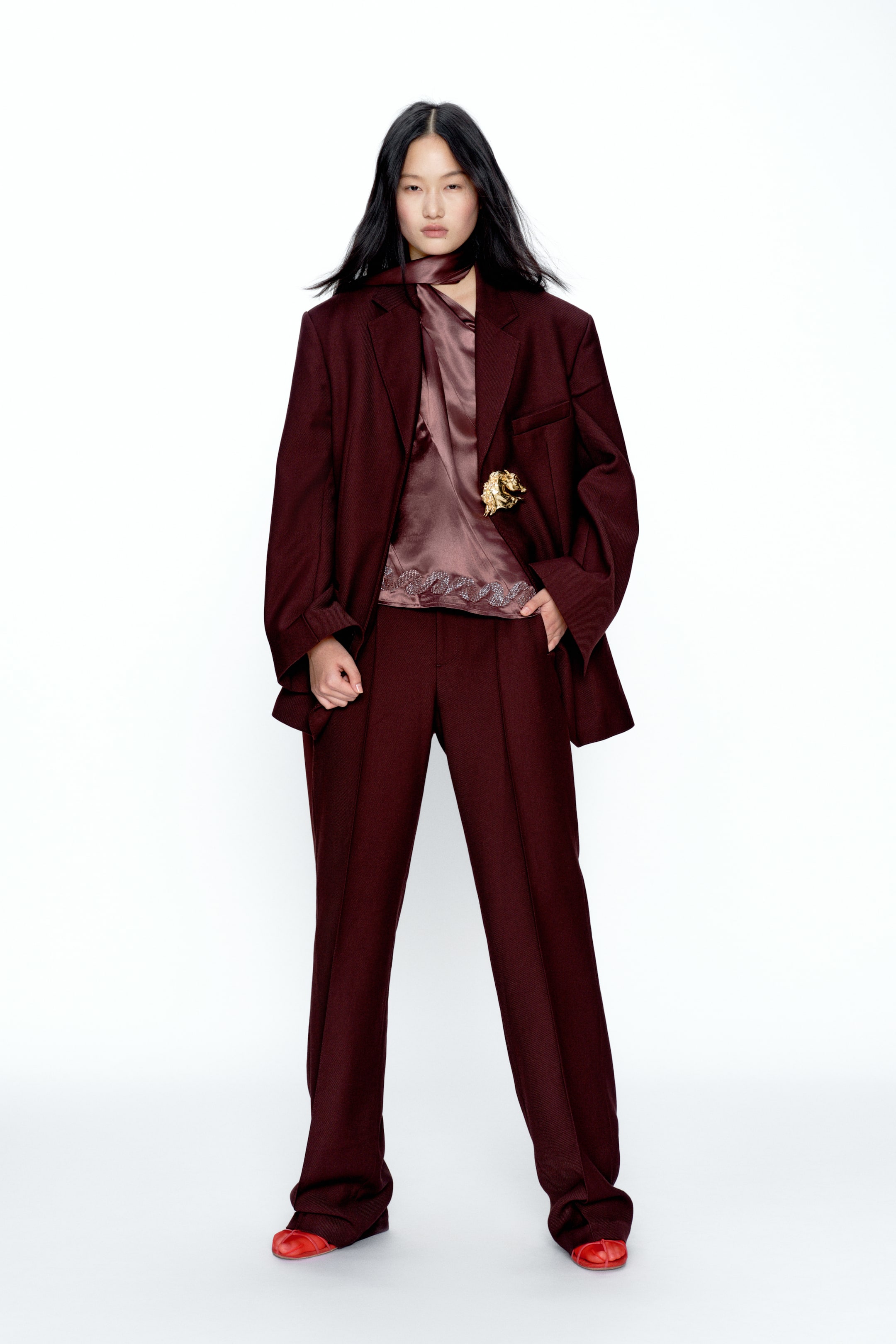 View larger image: A burgundy suit featuring an oversized blazer with notch lapels and wide-leg trousers with front creases, shown from the front. A light burgundy blouse with a tie-neck and lace trim is layered under the blazer, which also showcases a gold-toned brooch on its lapel.
