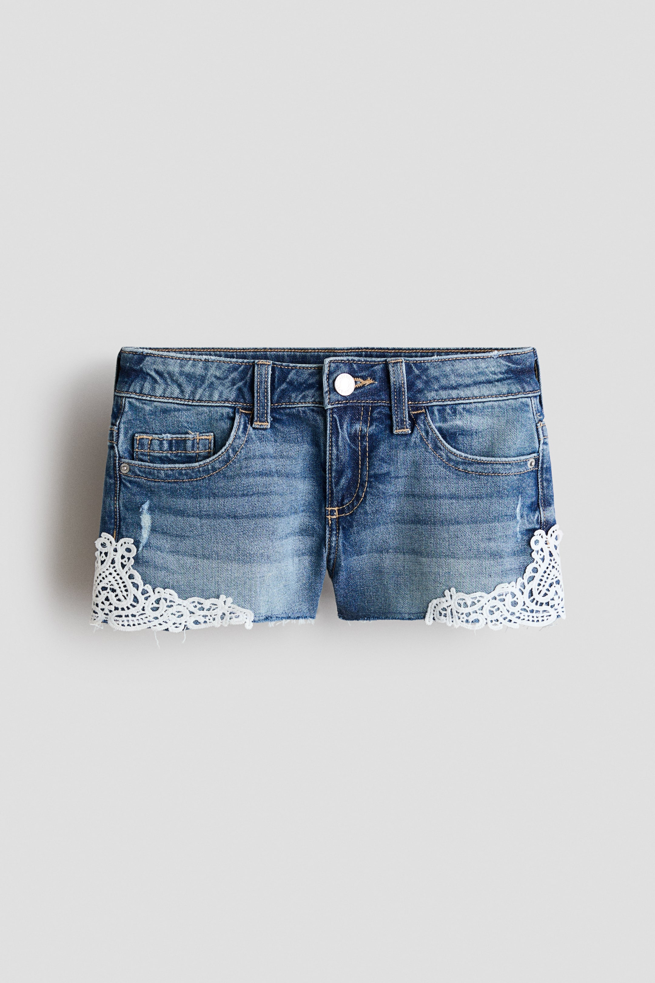 View larger image: Denim shorts showcase a medium blue, distressed wash with frayed raw hems. White lace trim embellishes the outer side seams of both legs, visible from a front angle.