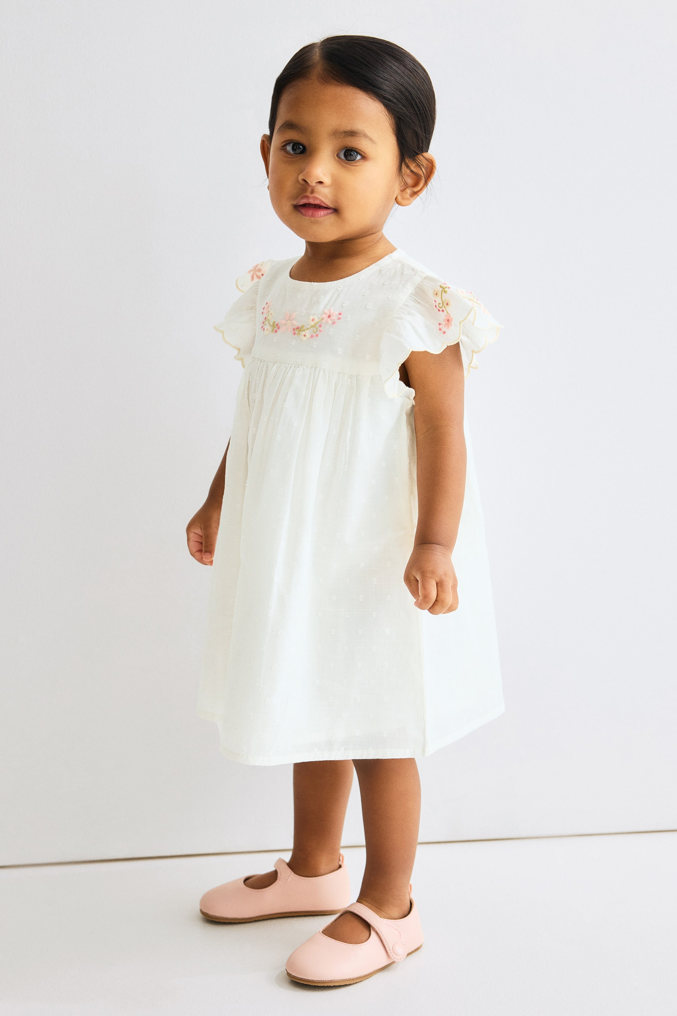 View larger image: A white frock crafted from a subtle dotted fabric showcases delicate pink and green floral embroidery adorning its round neckline and ruffled cap sleeves. The frock is gathered under the bodice, creating a flowing skirt that gracefully falls just above the knees, paired with light pink Mary Jane shoes.