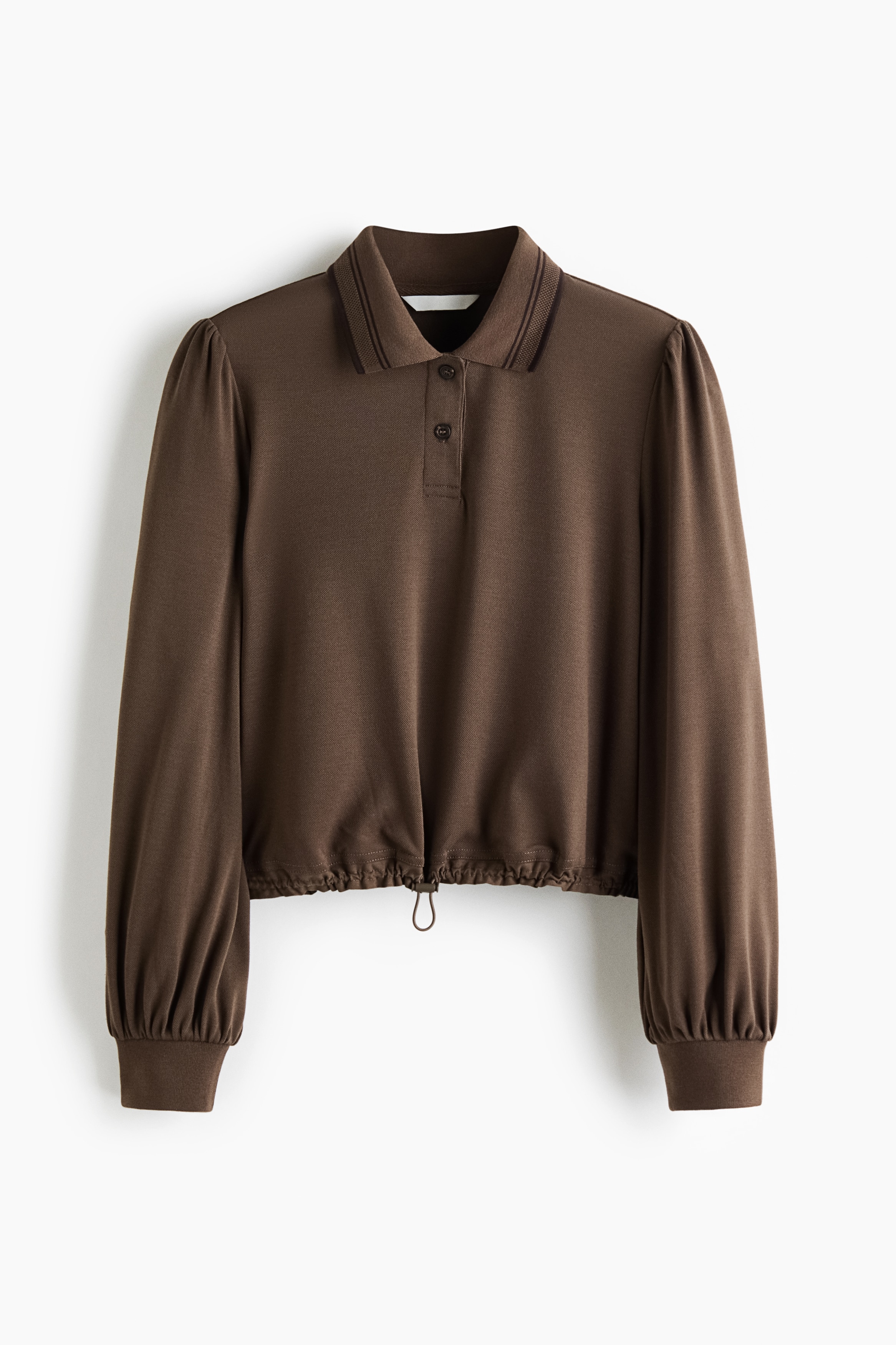 The model is wearing a brown polo shirt featuring a ribbed collar, button placket, long puff sleeves with ribbed cuffs, and a cropped drawstring hem with a cord stopper. The shirt is styled with light beige denim jeans.