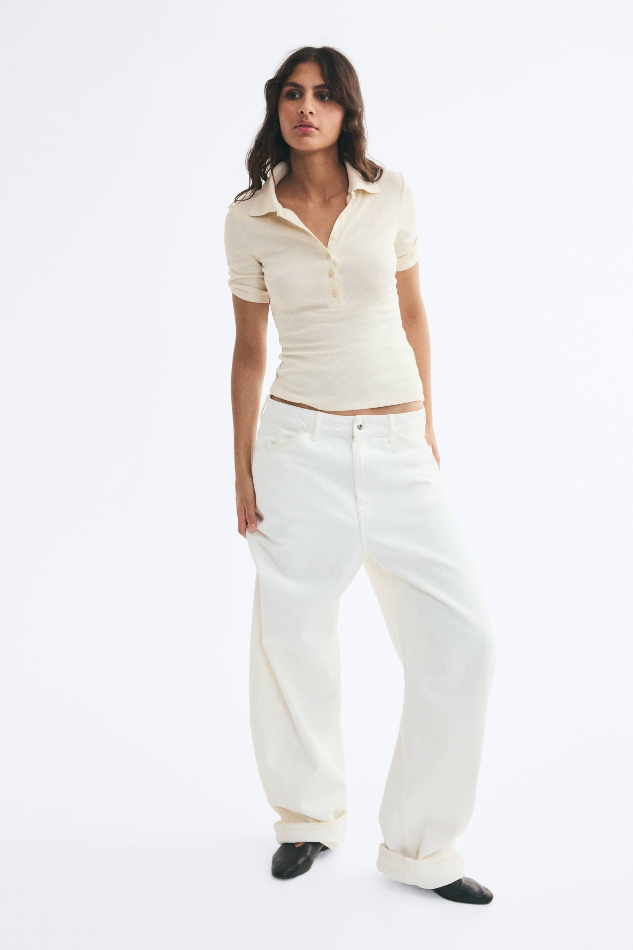 View larger image: A cream-colored, fitted polo shirt in a soft rib-knit fabric is being worn by the model. The shirt features a collar, a partially unbuttoned placket, and short sleeves, and is paired with wide-leg, off-white trousers.