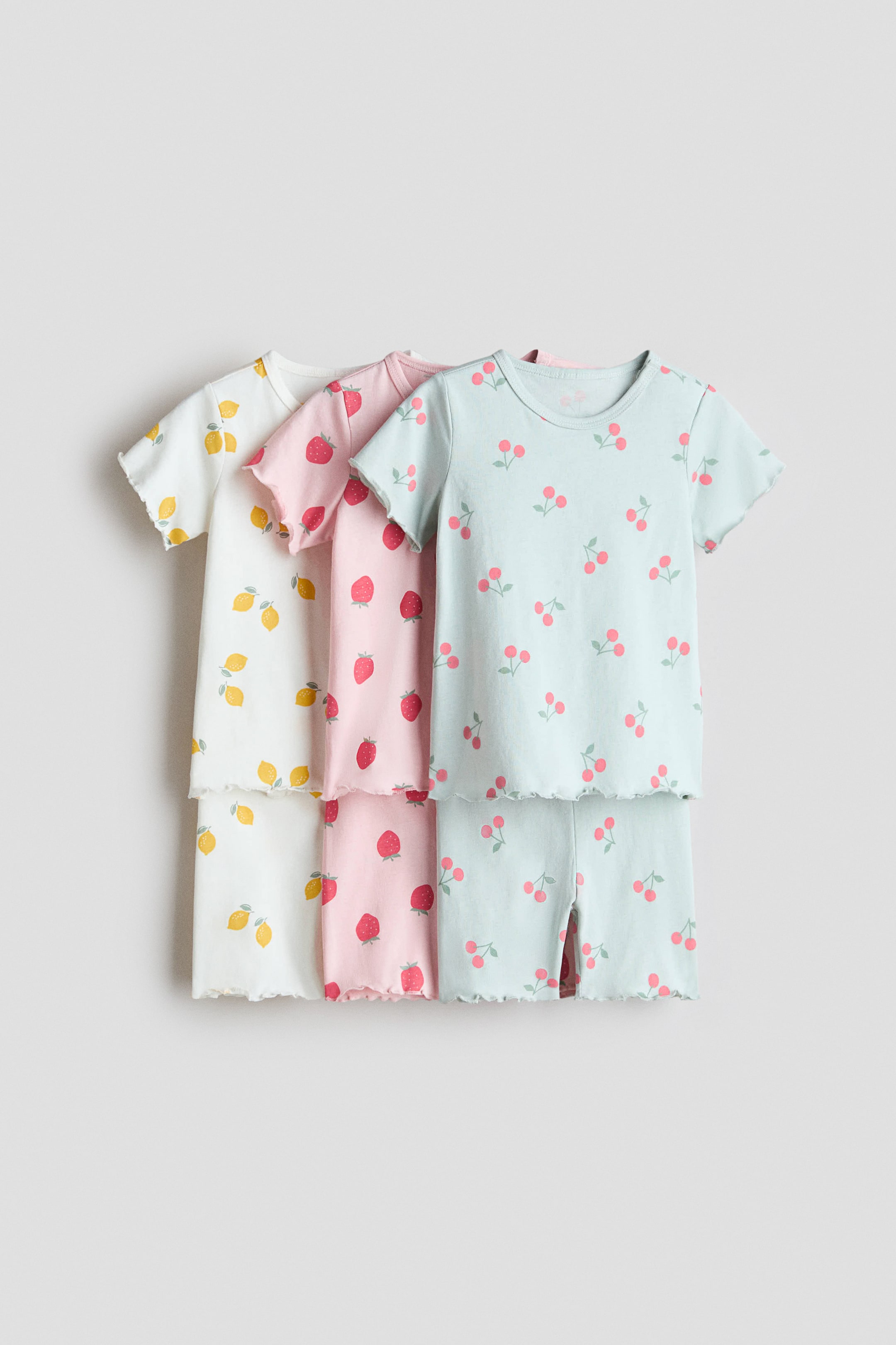 Agrandir l'image: Trois ensembles de pyjamas à manches courtes affichent leur vue de face. Chaque ensemble comprend un haut et un short assorti, et toutes les pièces présentent des bords à volants sur les manches et les ourlets. L'ensemble blanc montre un motif de citron, l'ensemble rose met en avant un motif de fraise, et l'ensemble bleu clair présente un motif de cerise.