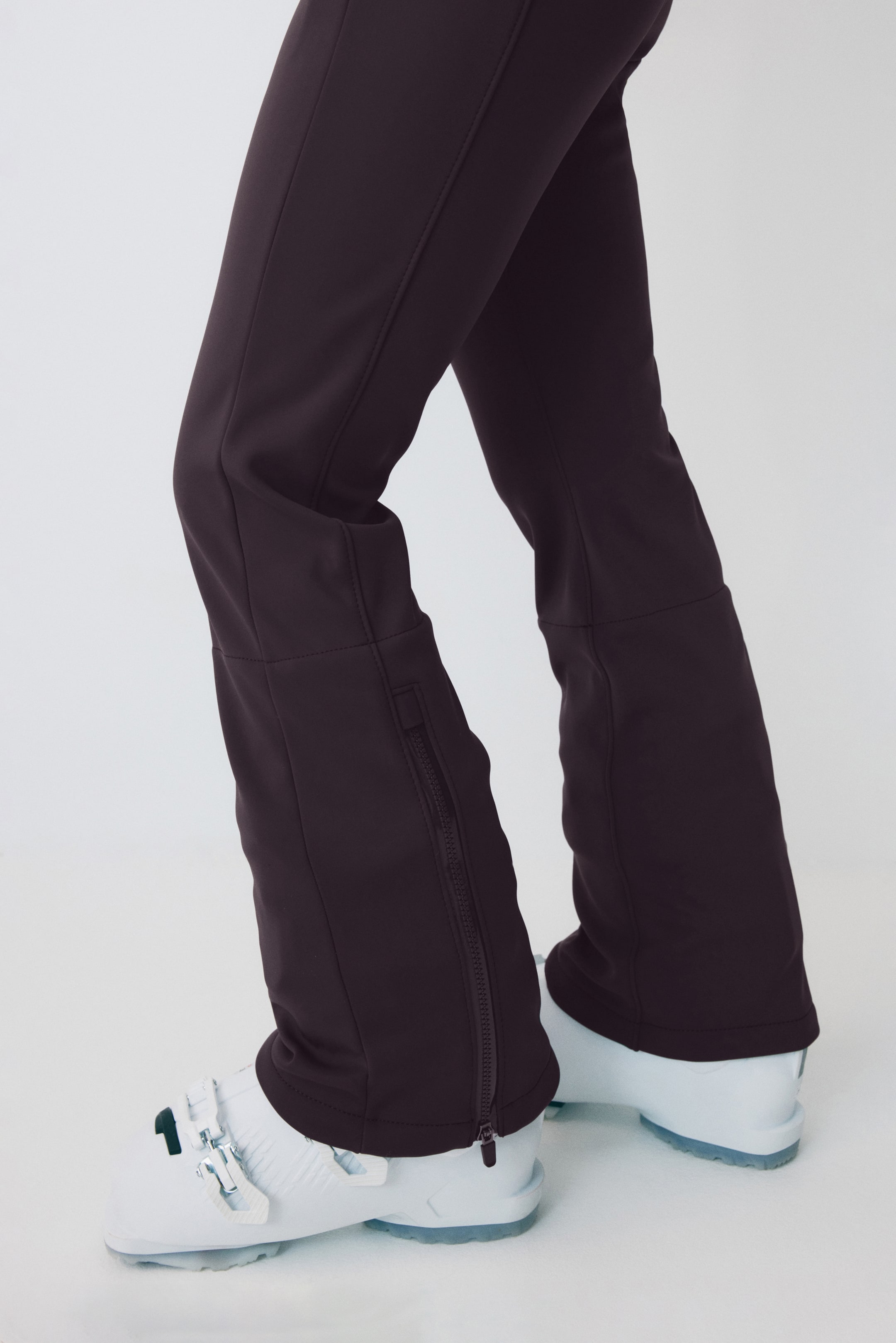 Agrandir l'image: Un pantalon de ski marron foncé s'ajuste parfaitement sur des bottes de ski blanches, avec des coutures d'articulation visibles aux genoux et des coutures verticales le long de l'avant des jambes. Chaque bas de jambe montre une fermeture éclair sombre sur son côté extérieur, s'étendant de la cheville vers le haut.