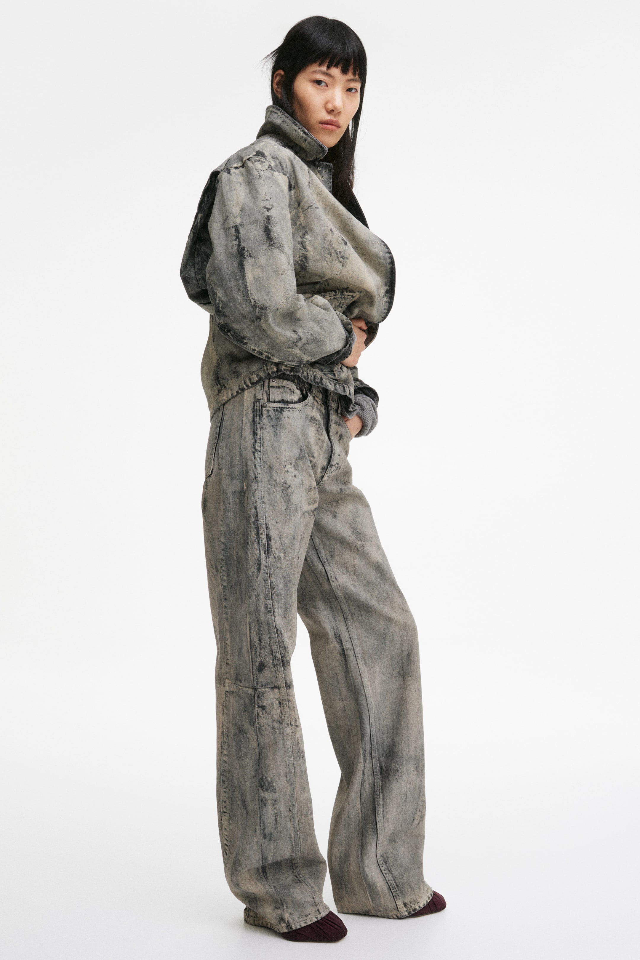 View larger image: "A coordinated acid wash denim jacket and wide-leg jeans are styled, shown from a slight right angle. The jacket showcases a high collar and dropped shoulders, with a grey ribbed cuff peeking out from beneath. Meanwhile, the high-waisted jeans offer a relaxed fit, elegantly gathering over dark red socks."