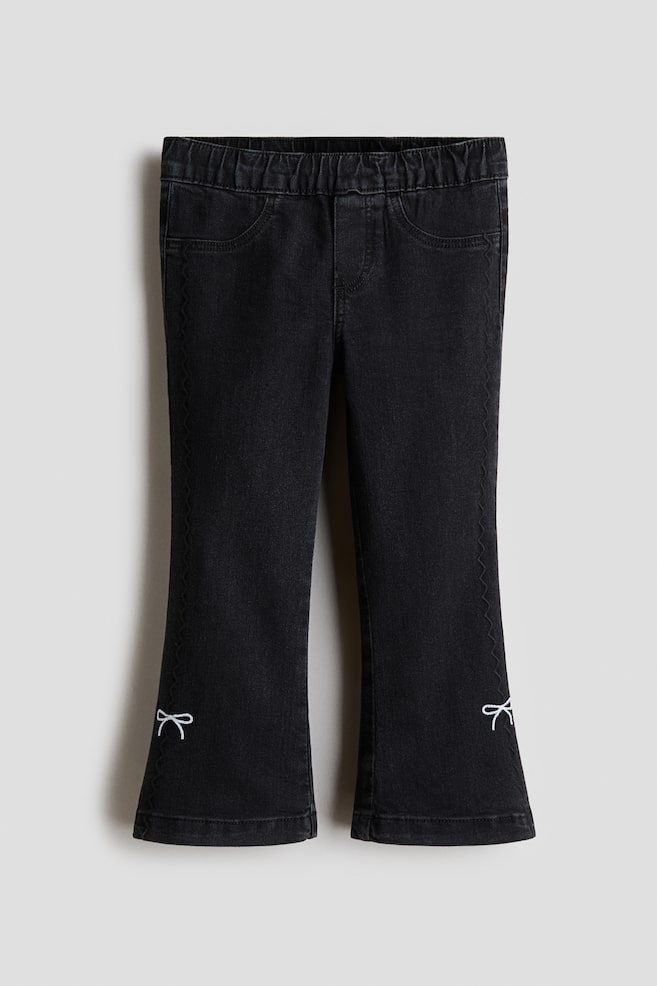 Black children's pants feature an elasticated waistband, faux front pockets, and decorative zig-zag stitching along the outer seams, with white embroidered bows adorning the flared lower legs. - 1