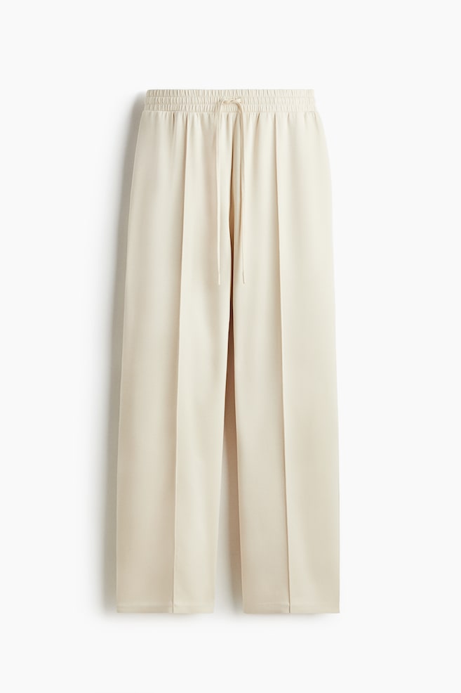 Front view of light beige wide-leg pants, featuring an elasticized, gathered waistband with a visible drawstring. Vertical creases are running down the front of each leg. - 1