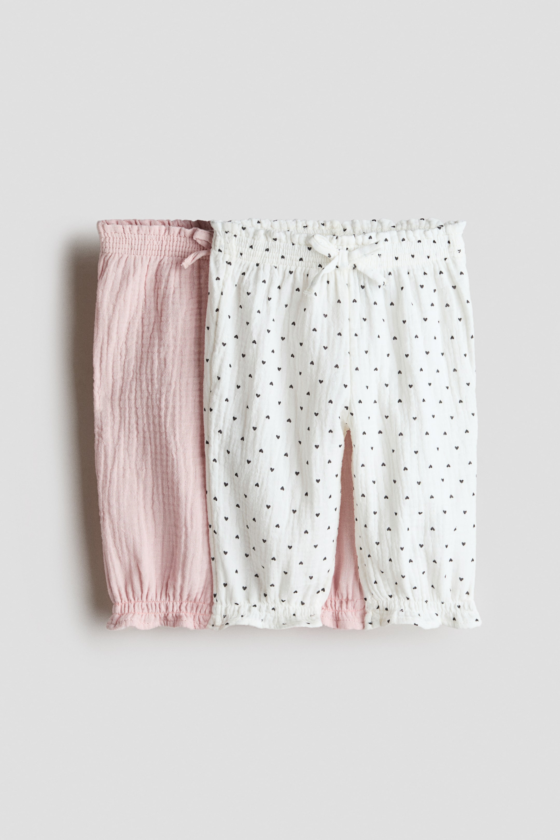 View larger image: Two pairs of trousers in soft cotton muslin are showcasing their front views. One pair is light pink, while the other is white adorned with a small black heart pattern. Both feature a decorative bow at the front, smocked elastication at the waist, and frill trim with smocked elastication at the hems.