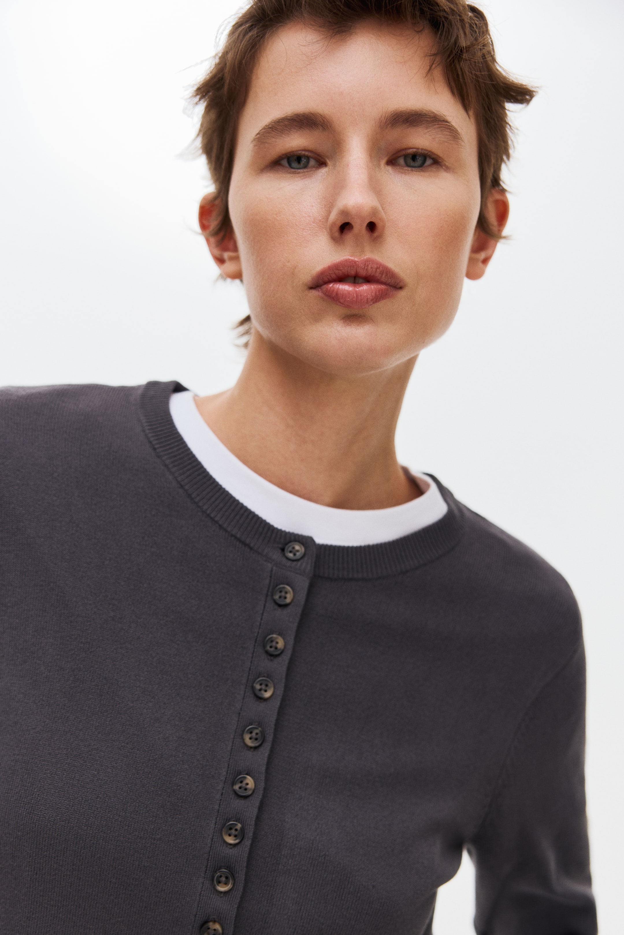 View larger image: A dark grey knit cardigan is worn over a white T-shirt, featuring a ribbed crew-neck and a front button placket with small brown buttons.