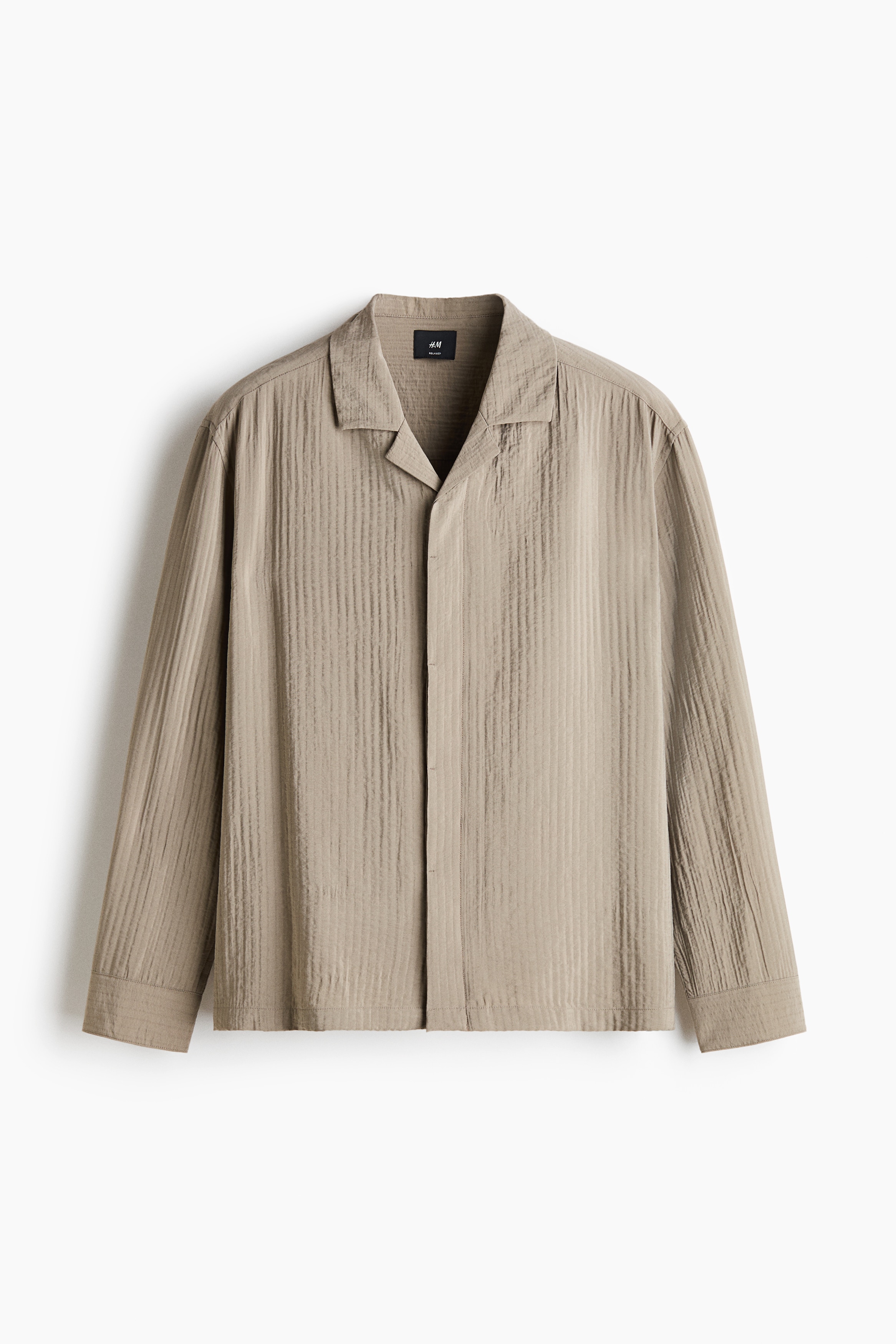 A light beige, textured-weave long-sleeve shirt with a resort collar is being worn unbuttoned by the model. The shirt is layered over a white ribbed tank top and paired with off-white trousers.