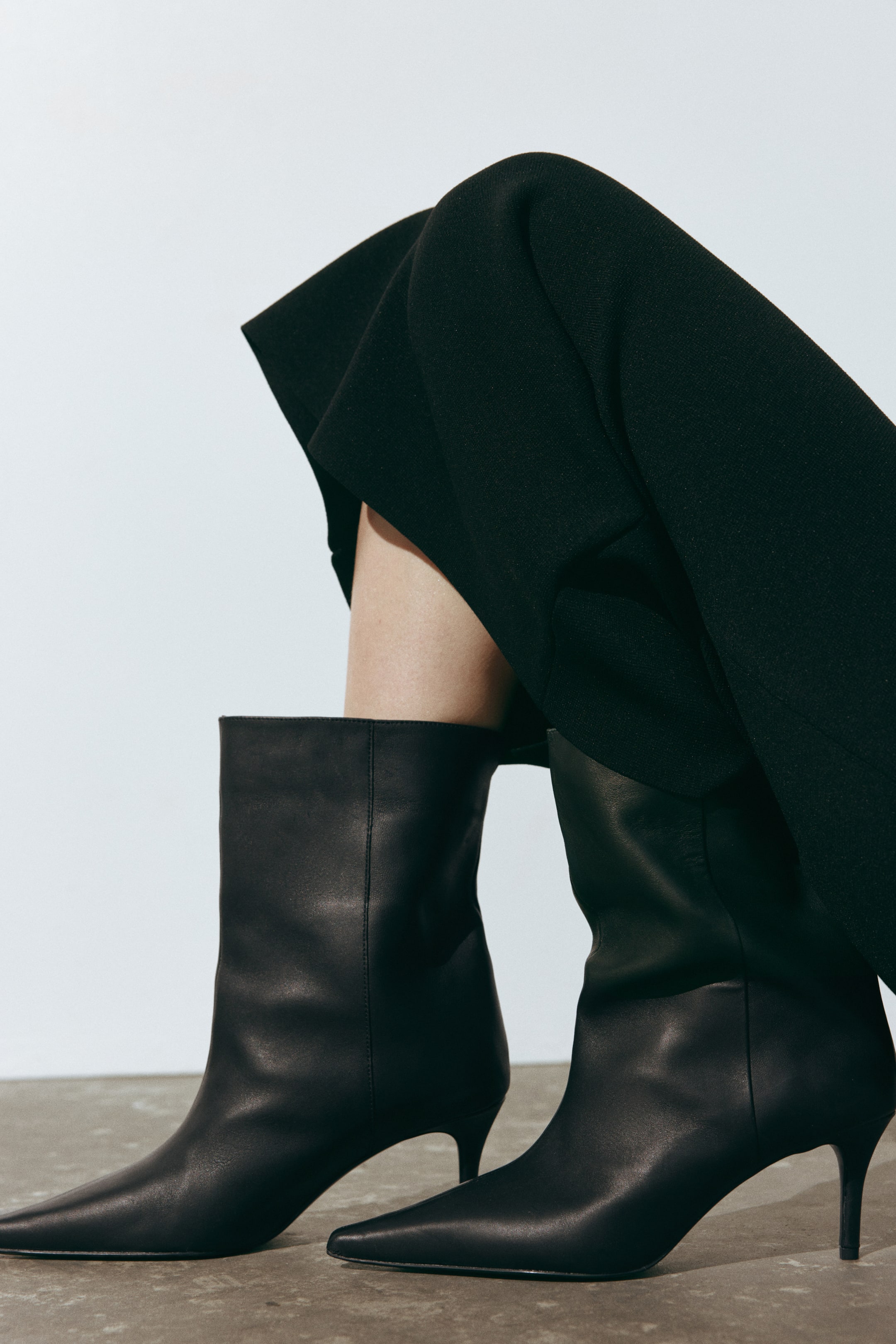Agrandir l'image: Des bottes en cuir noir à tiges larges arrivant à la cheville, à bouts pointus et à talons fins recouverts sont portées par le mannequin. Les bottes sont associées à une jupe noire drapée qui retombe sur les jambes, mettant en valeur la largeur des tiges.