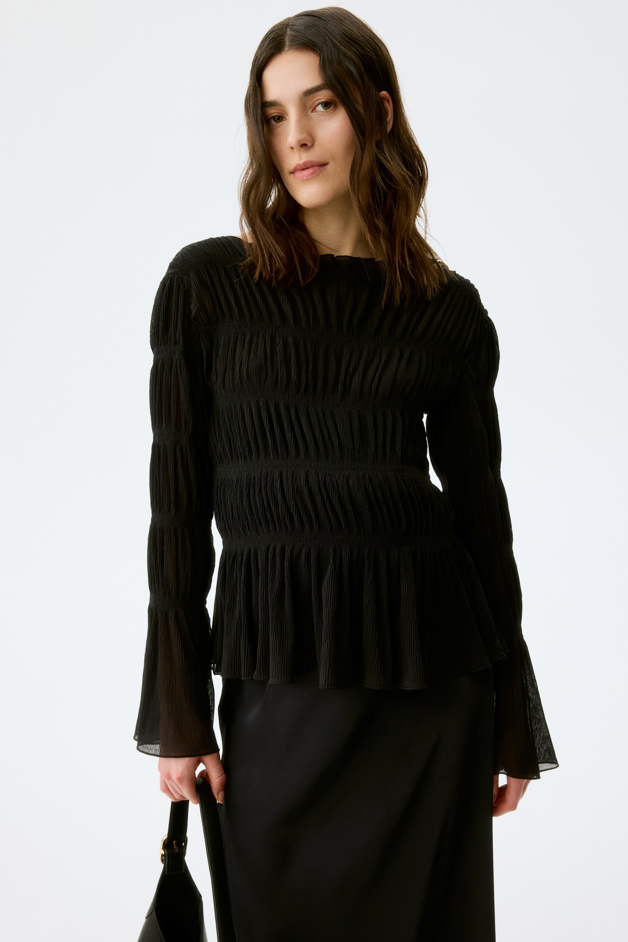 Ver imagem maior: O top preto apresenta um decote em barco e um corpete franzido com uma textura enrugada, estendendo-se em mangas compridas com punhos transparentes e alargados. A bainha em peplum abre-se na cintura, usada sobre uma saia preta.