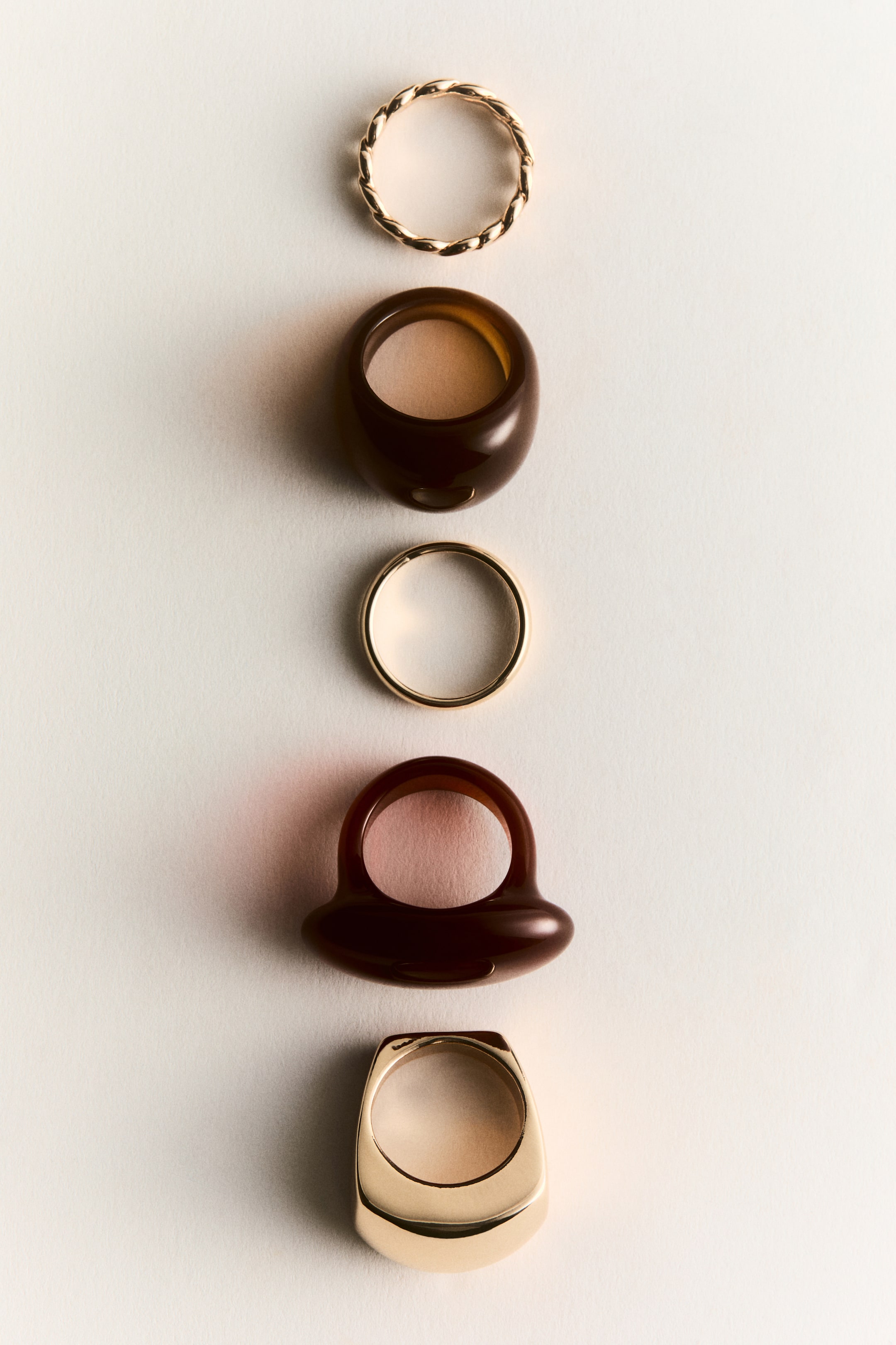 Zobrazit větší obrázek: Pět prstenů je uspořádáno vertikálně, každý s odlišným designem a materiálem. Horní prsten je zkroucený prsten s odstínem zlata, následuje silný, zaoblený, tmavě hnědý průsvitný prsten. Dále je umístěn jednoduchý, tenký prsten s odstínem zlata, poté tmavě hnědý průsvitný prsten se širokým, zaobleným, zploštělým vrškem, a masivní prsten s pečetním kamenem s plochým, čtvercovým vrškem dokončuje uspořádání.