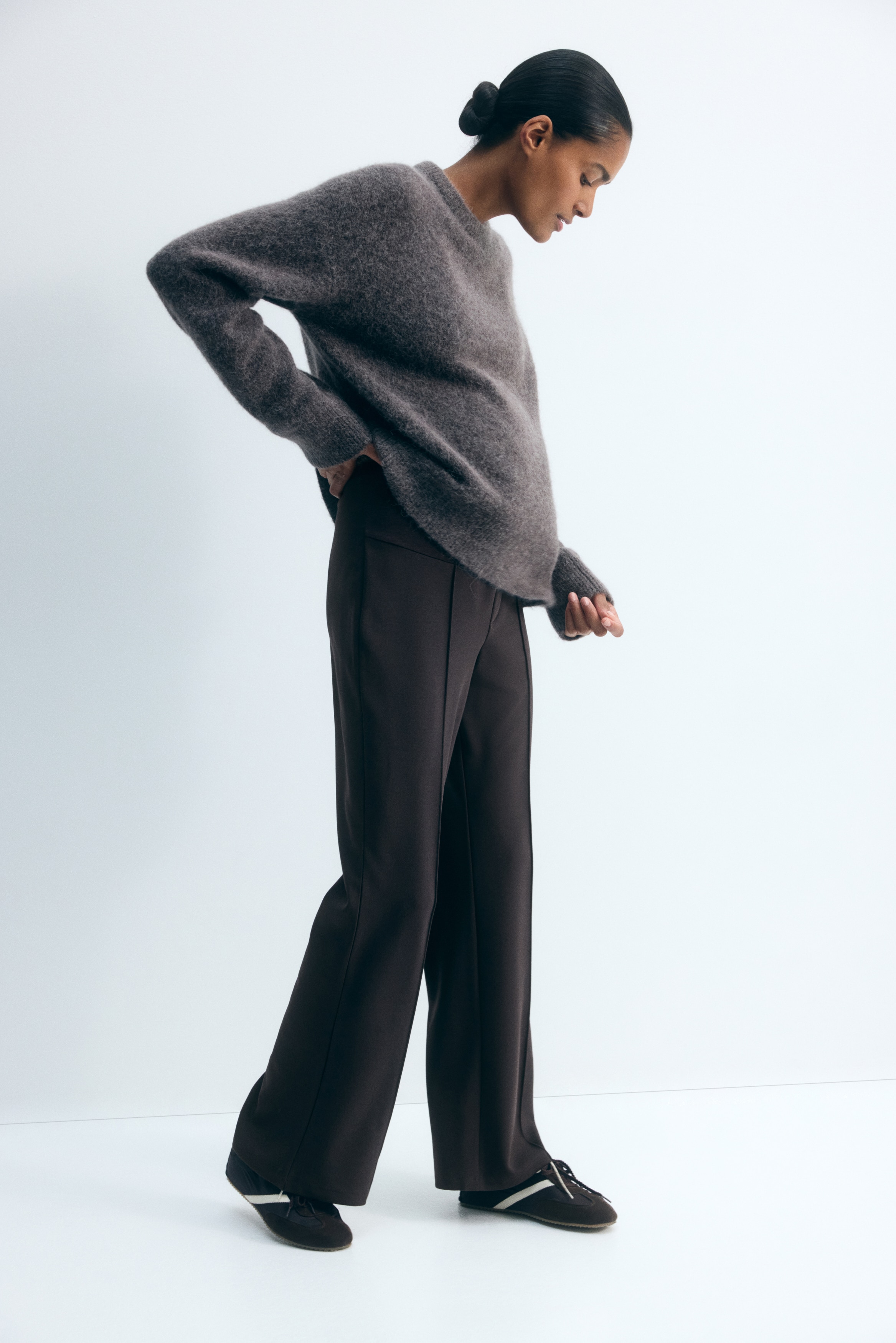 Black maternity trousers are shown from the front, featuring a high, elasticated over-the-bump waistband and a straight-leg silhouette. The trousers display sharp pressed creases running down the centre of each leg and a faux fly detail below the band.