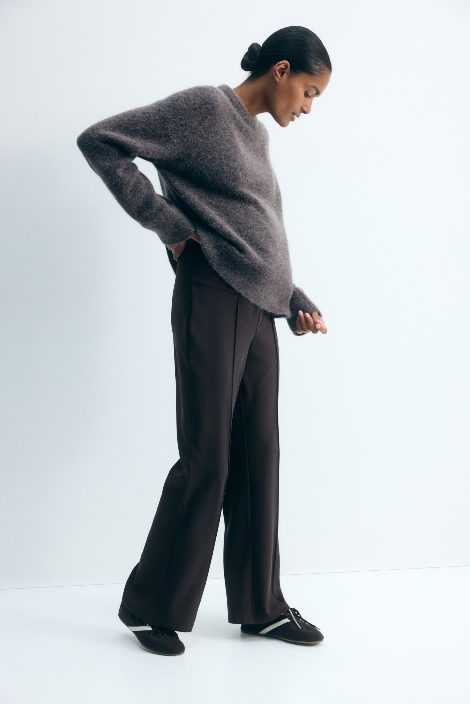 Dark brown, bootcut jersey trousers featuring sewn-in creases down the front are worn by the model. The trousers have a high-rise waist with a wide waistband, which the model is showcasing by resting a hand on her hip beneath a loose grey knit jumper. - 1