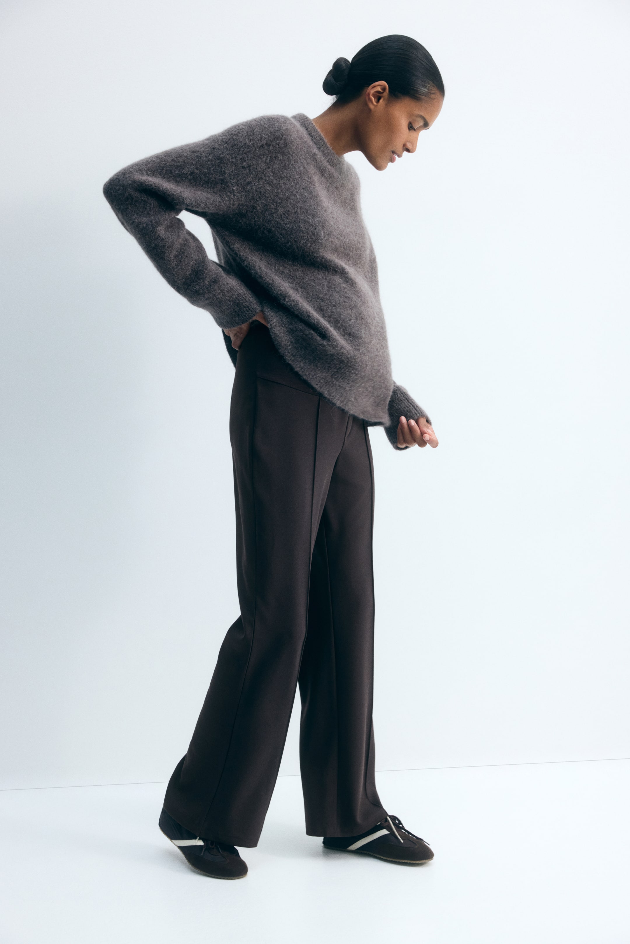Se større billede: Mørkebrune bootcut-jerseybukser med syede pressefolder ned langs forsiden bæres af modellen. Bukserne har en høj talje med et bredt panel, som modellen fremhæver ved at hvile en hånd på hoften under en løs grå striktrøje.