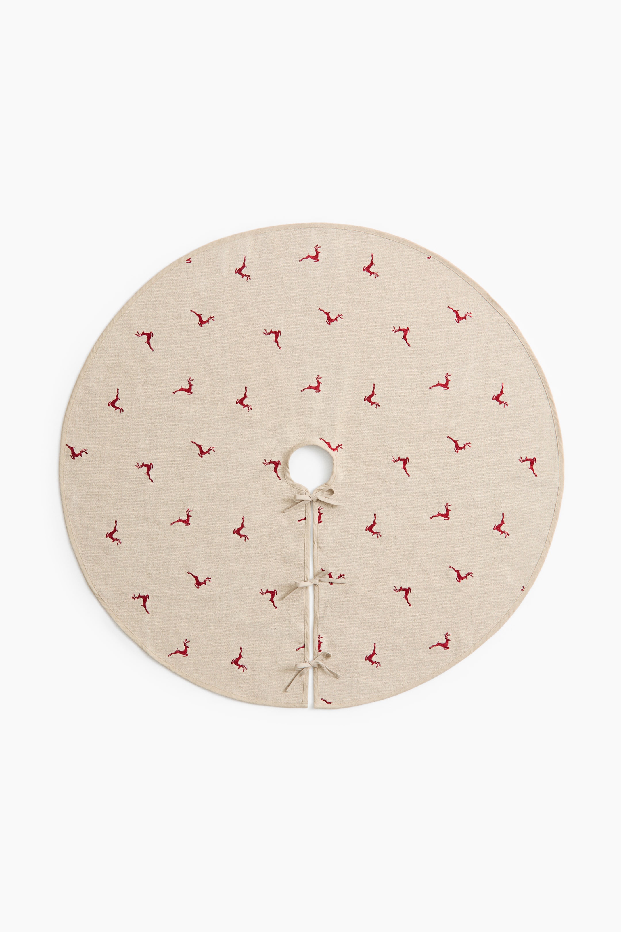 View larger image: A circular beige tree skirt displays a scattered pattern of small red reindeer. It features a central opening and a straight slit secured by three matching fabric ties tied in bows.