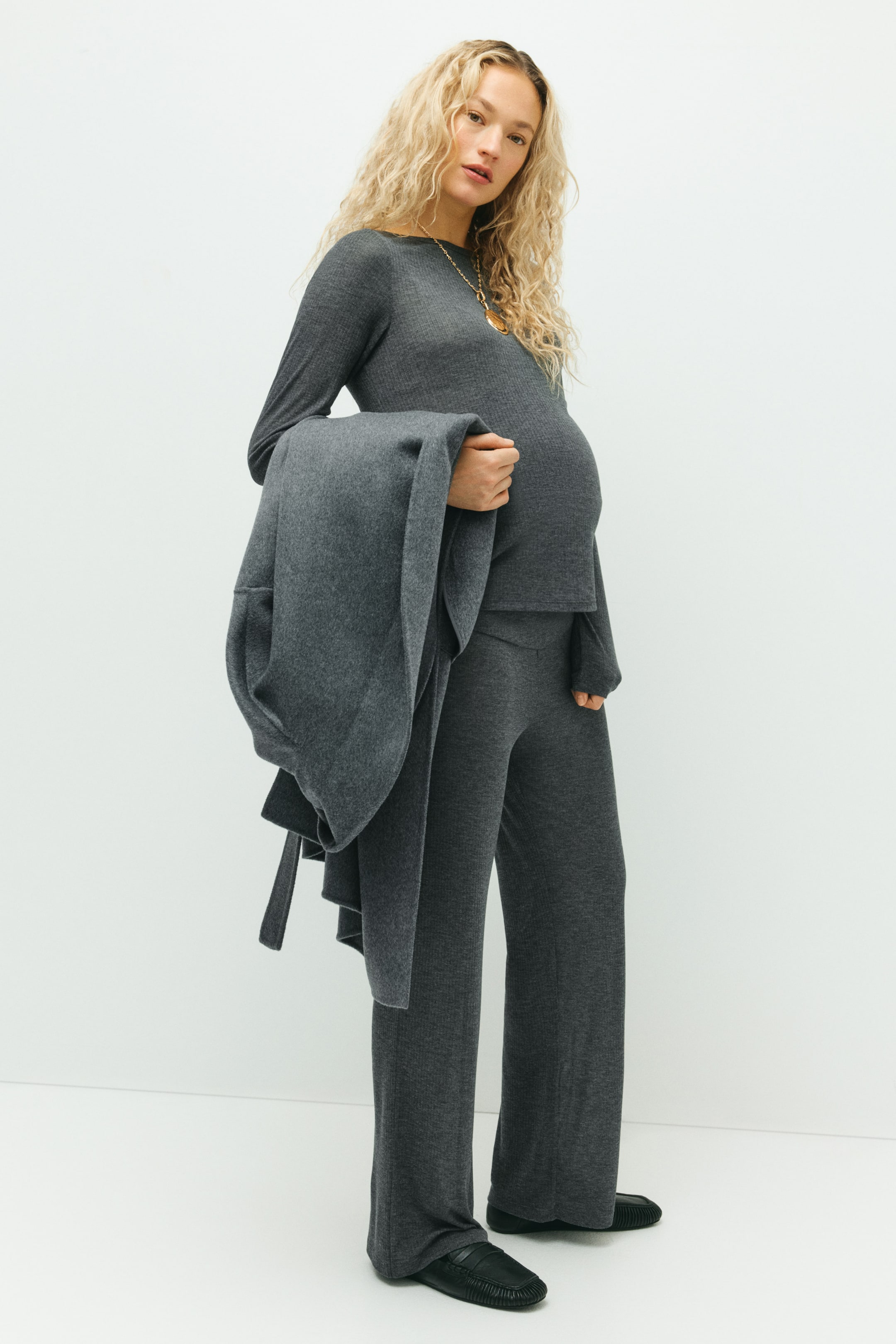 Vis større bilde: Et mørkegrått ribbestrikket jerseyssett, med en langermet topp med båthals og bukser med rette ben, bæres av modellen. Buksene passer over modellens gravide mage, og hun holder et matchende grått plagg over armen.