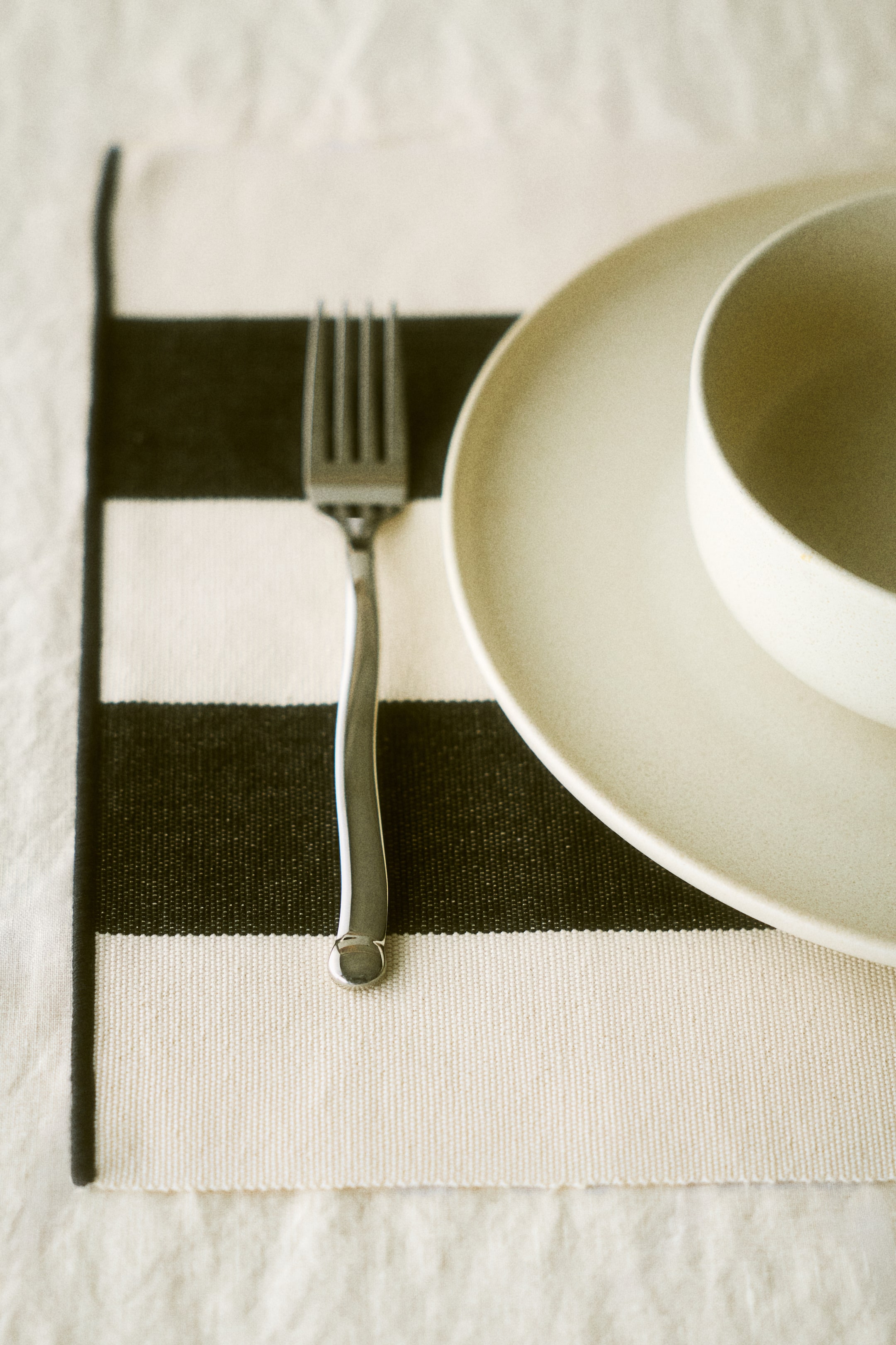 View larger image: A woven placemat showcases alternating horizontal black and off-white stripes. A silver-toned fork is placed on the left side of the placemat, while a light beige round dinner plate holds a nested bowl on the right.