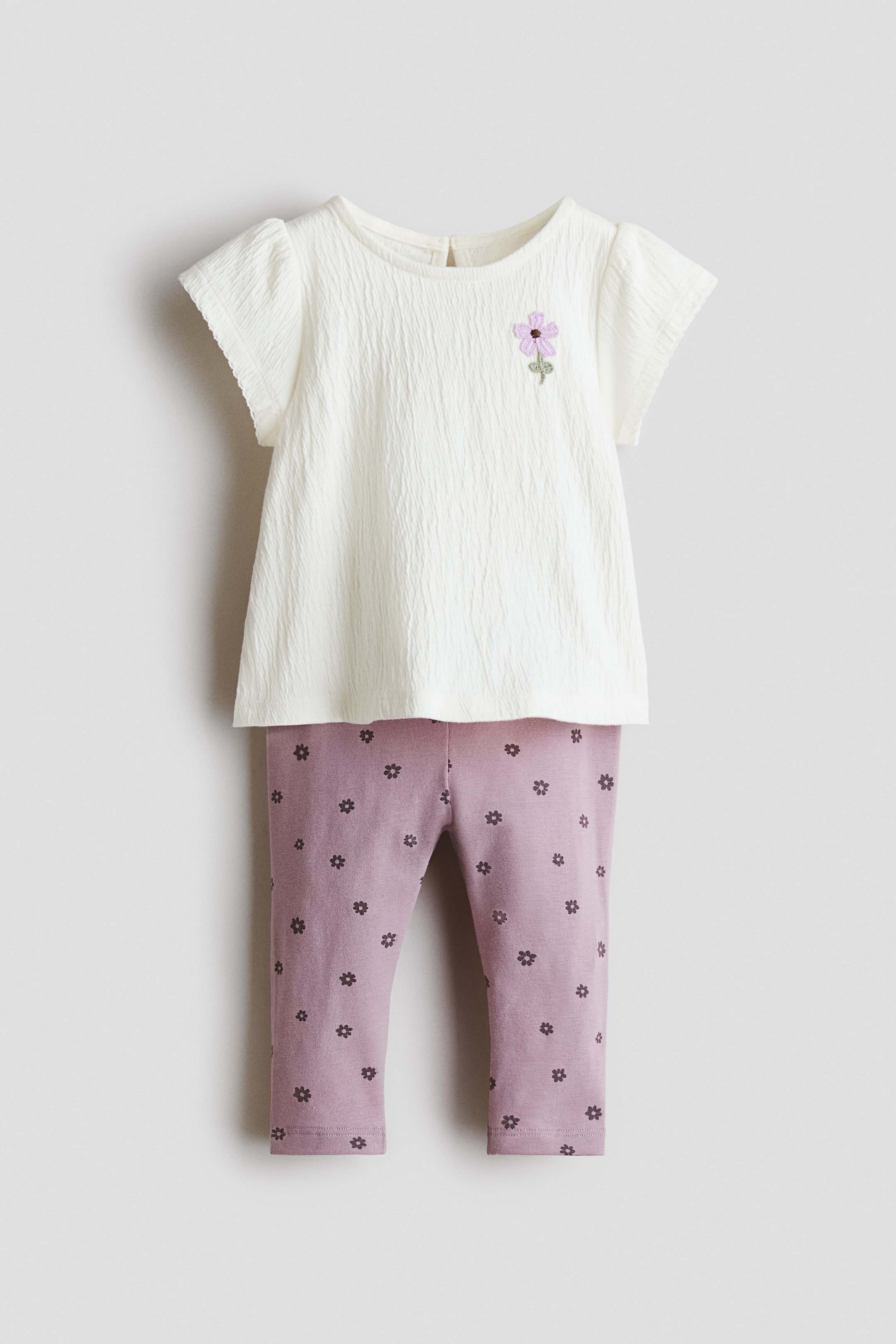View larger image: A baby’s outfit features an off-white textured top with cap sleeves, a back button closure, and a purple embroidered flower on the chest, layered over mauve leggings patterned with small dark floral motifs.