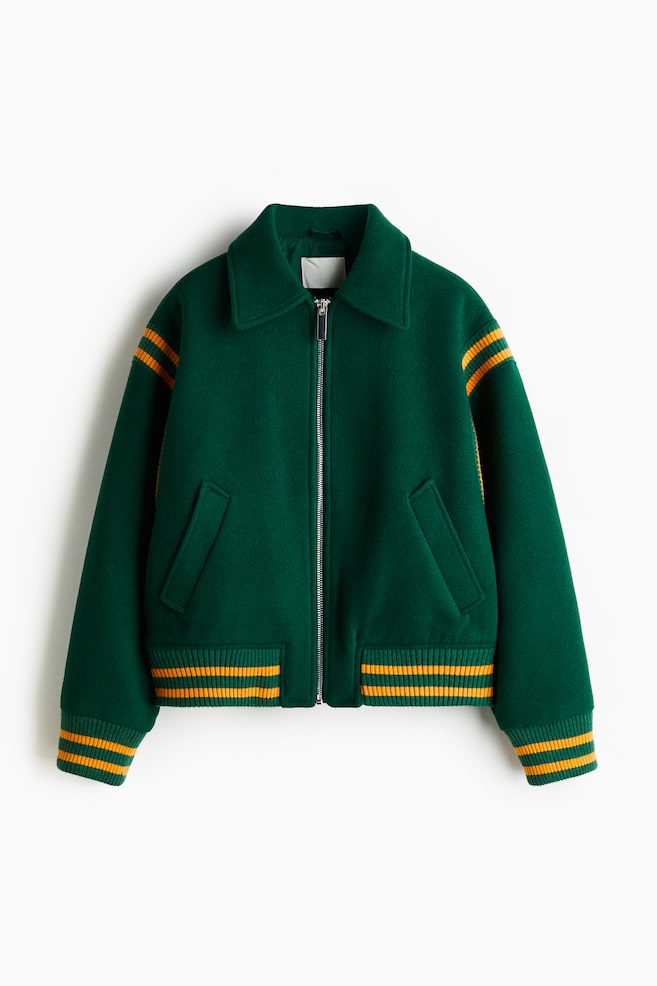 Eine dunkelgrüne Jacke wird von vorne präsentiert, mit einem silberfarbenen Reißverschluss in voller Länge, einem spitzen Kragen und zwei schrägen Leistentaschen. Sie ist mit gerippten Bündchen und einem gerippten Bund versehen, zusammen mit den oberen Ärmeln, die alle zwei horizontale gelbe Streifen zeigen. - 1
