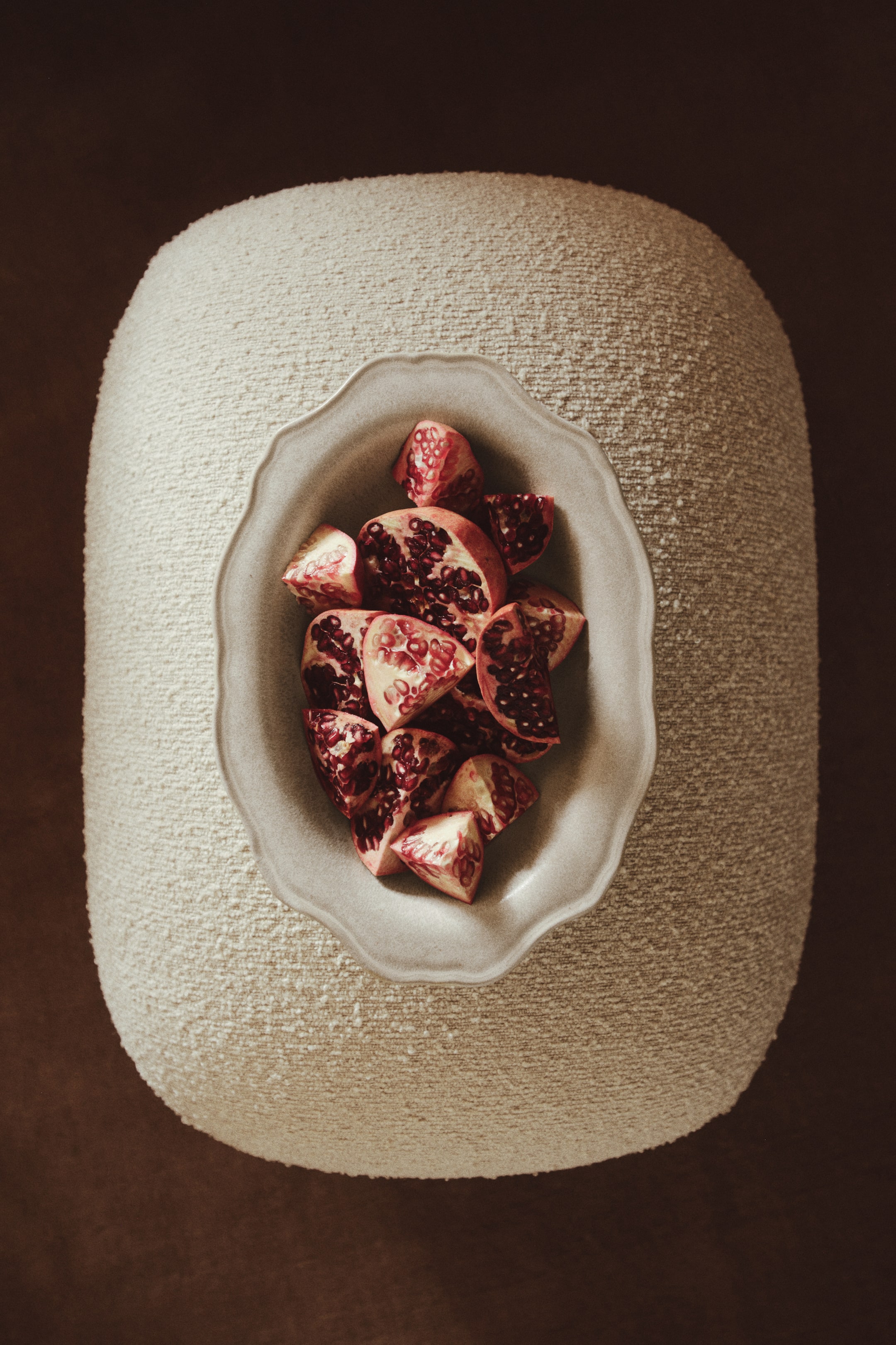 View larger image: A rounded, cream-coloured bouclé ottoman is shown from a top-down perspective, displaying a highly textured, nubby surface. An oval ceramic bowl with a scalloped rim rests at the centre of the ottoman, holding several cut pieces of pomegranate.