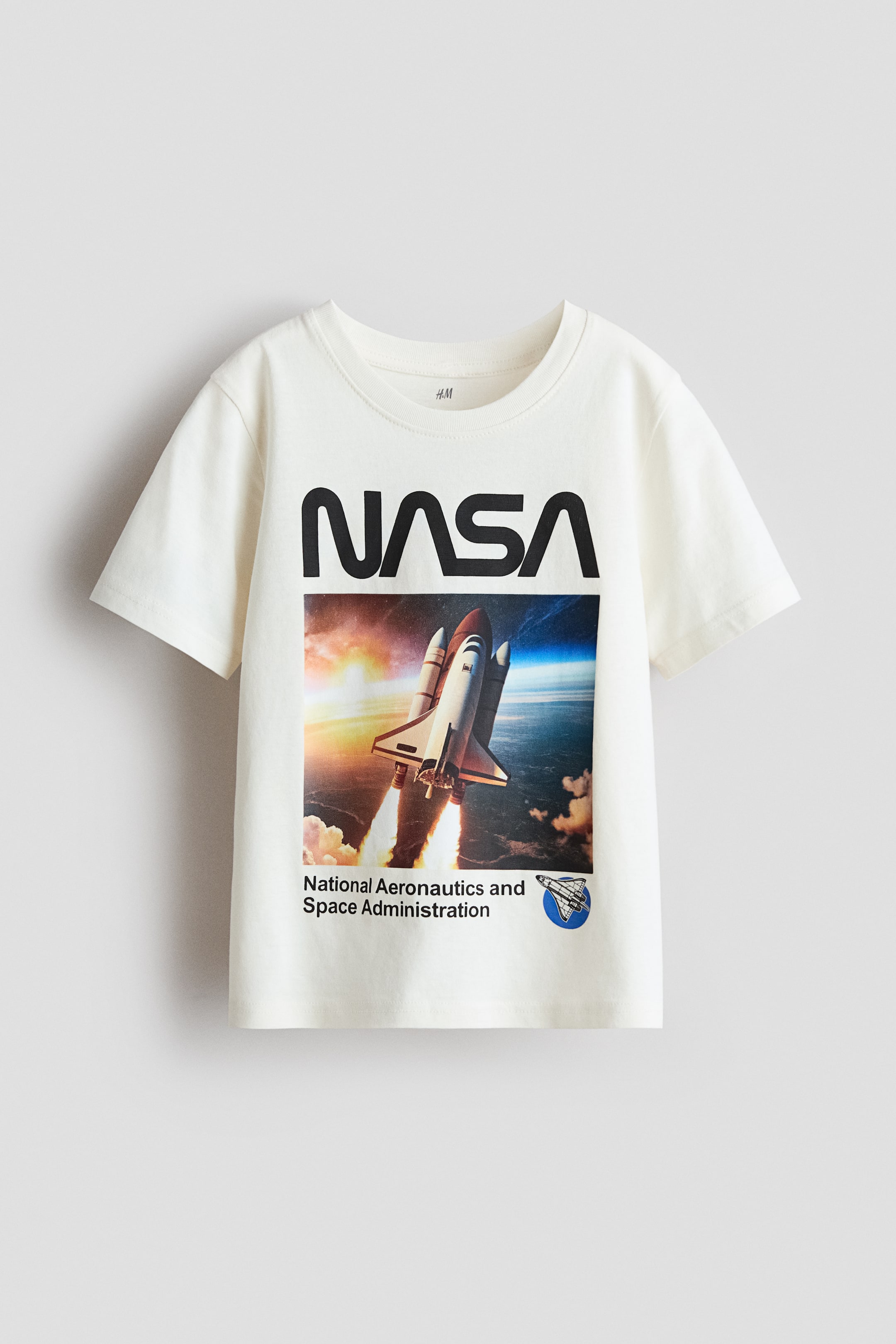 View larger image: A soft, off-white cotton jersey T-shirt is shown from the front, featuring a round, ribbed neckline and slightly dropped shoulders. A large graphic print covers the chest, displaying "NASA" in black block letters above an image of a space shuttle launch, with "National Aeronautics and Space Administration" and a small space shuttle logo printed below.