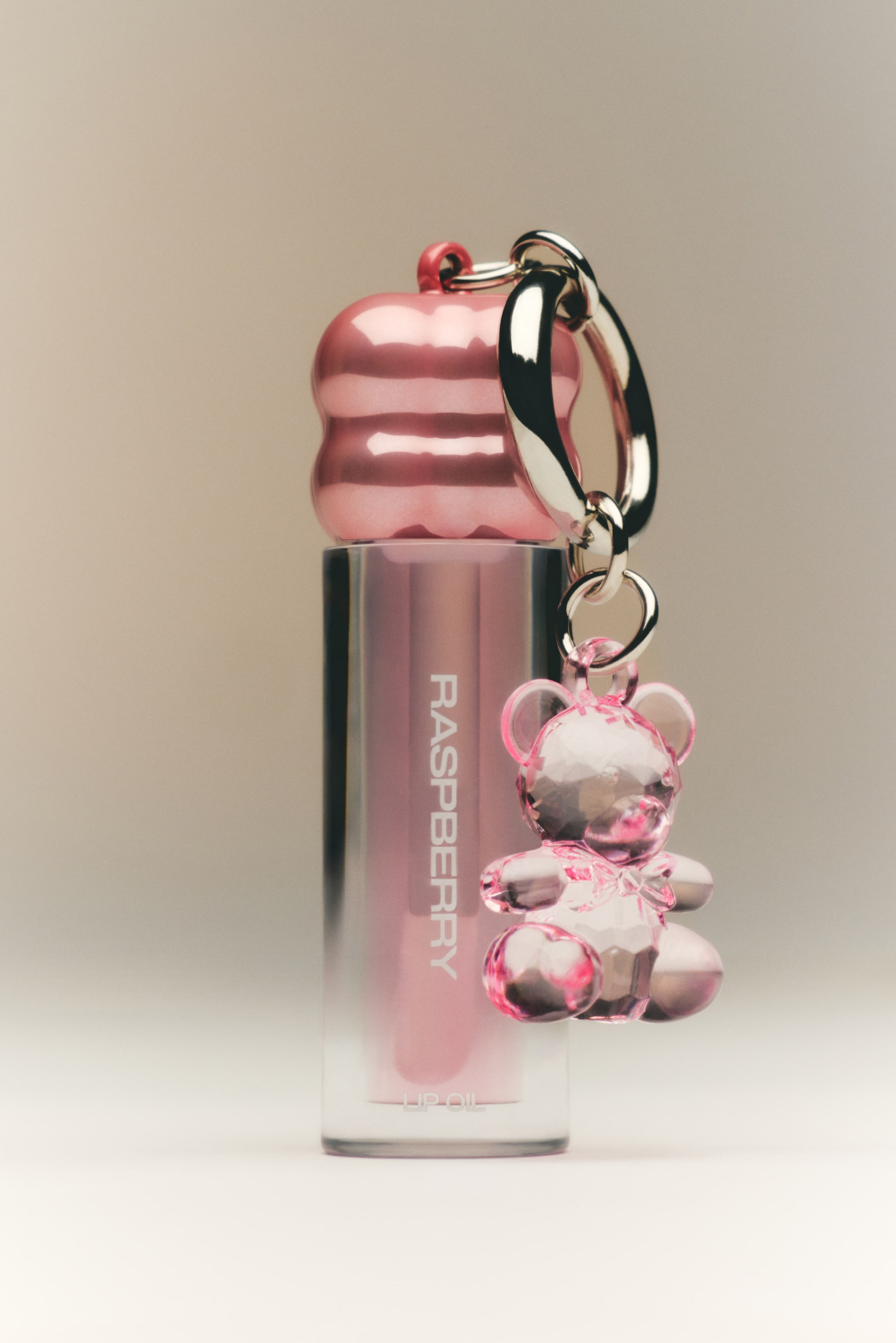 Grotere afbeelding bekijken: Een rechtopstaande sleutelhanger toont een lipolie in een heldere cilindrische tube met een roze vloeistof waarop het etiket 'RASPBERRY LIP OIL' staat. De tube wordt afgesloten door een glanzende roze geribbelde dop, en aan de dop is een zilverkleurige sleutelring met schakels bevestigd, waaraan een gefacetteerde, doorschijnend roze beertjesbedel hangt.