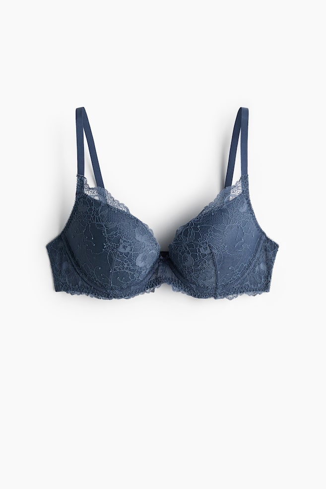 A deep blue push-up bra is laid flat, showcasing a lace overlay on its cups and scalloped lace trimming the top and bottom edges. A small bow adorns the centre gore, and thin adjustable straps extend upwards. - 1