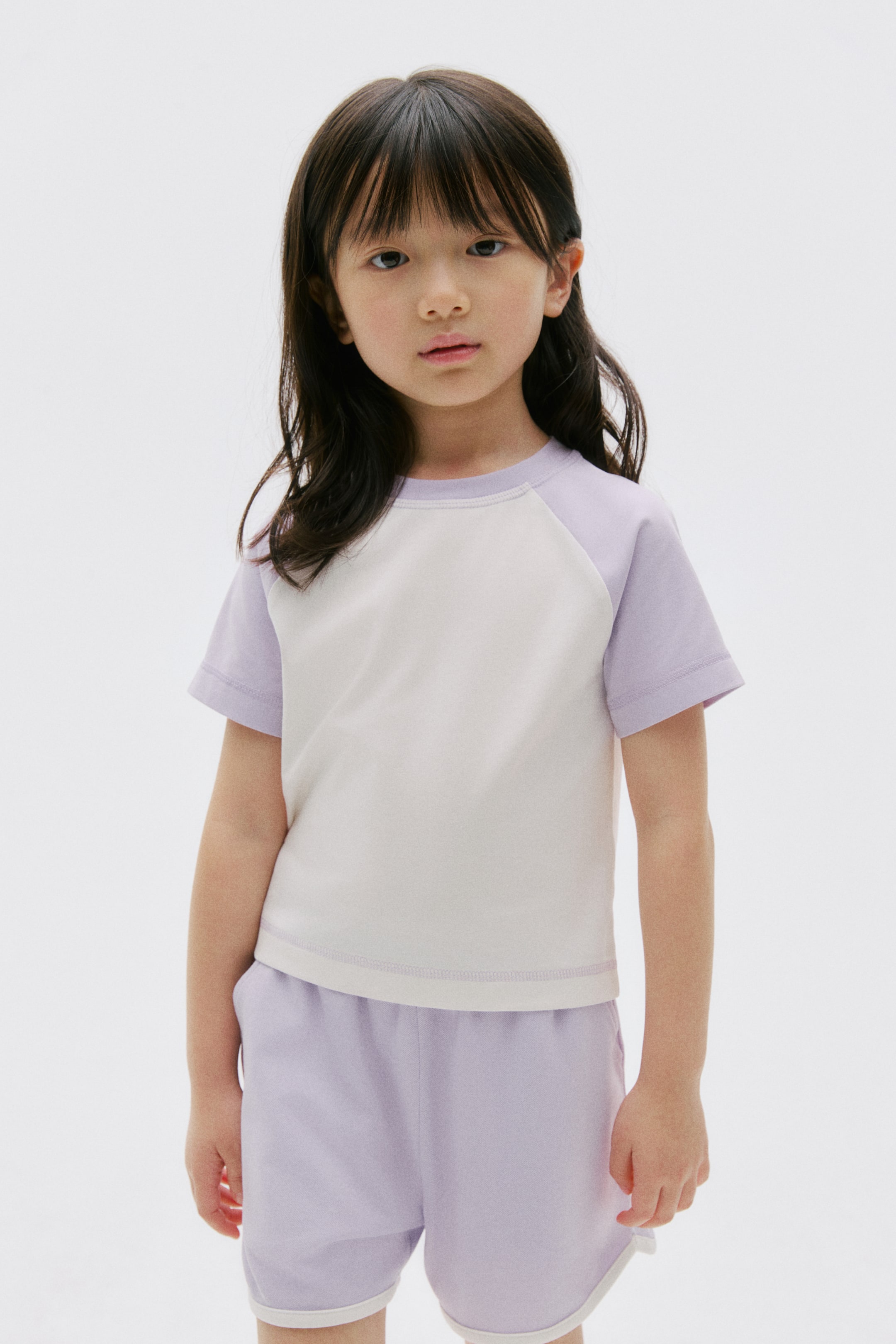 View larger image: The product is a two-piece set, featuring a light beige t-shirt with light purple raglan sleeves and a round neck, paired with matching light purple shorts. The shorts are designed with an elastic waistband and white trim along the leg openings, and the t-shirt is showing a visible double-stitched hem.