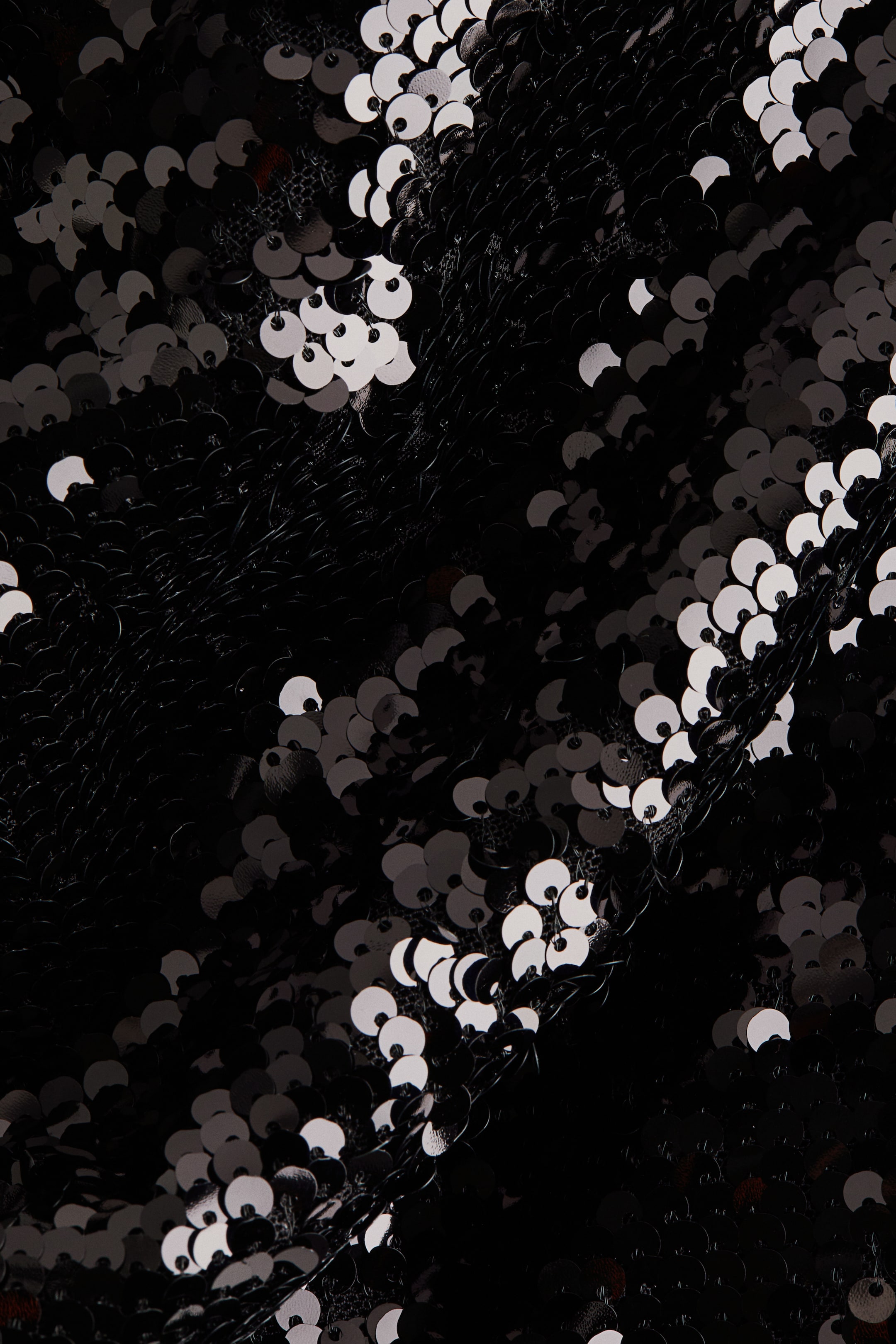 Visa större bild: Svart paljettyg är täckt med små, cirkulära svarta paljetter, där vissa reflekterar ljus som vitt eller silver, vilket skapar en texturerad och vågig yta.