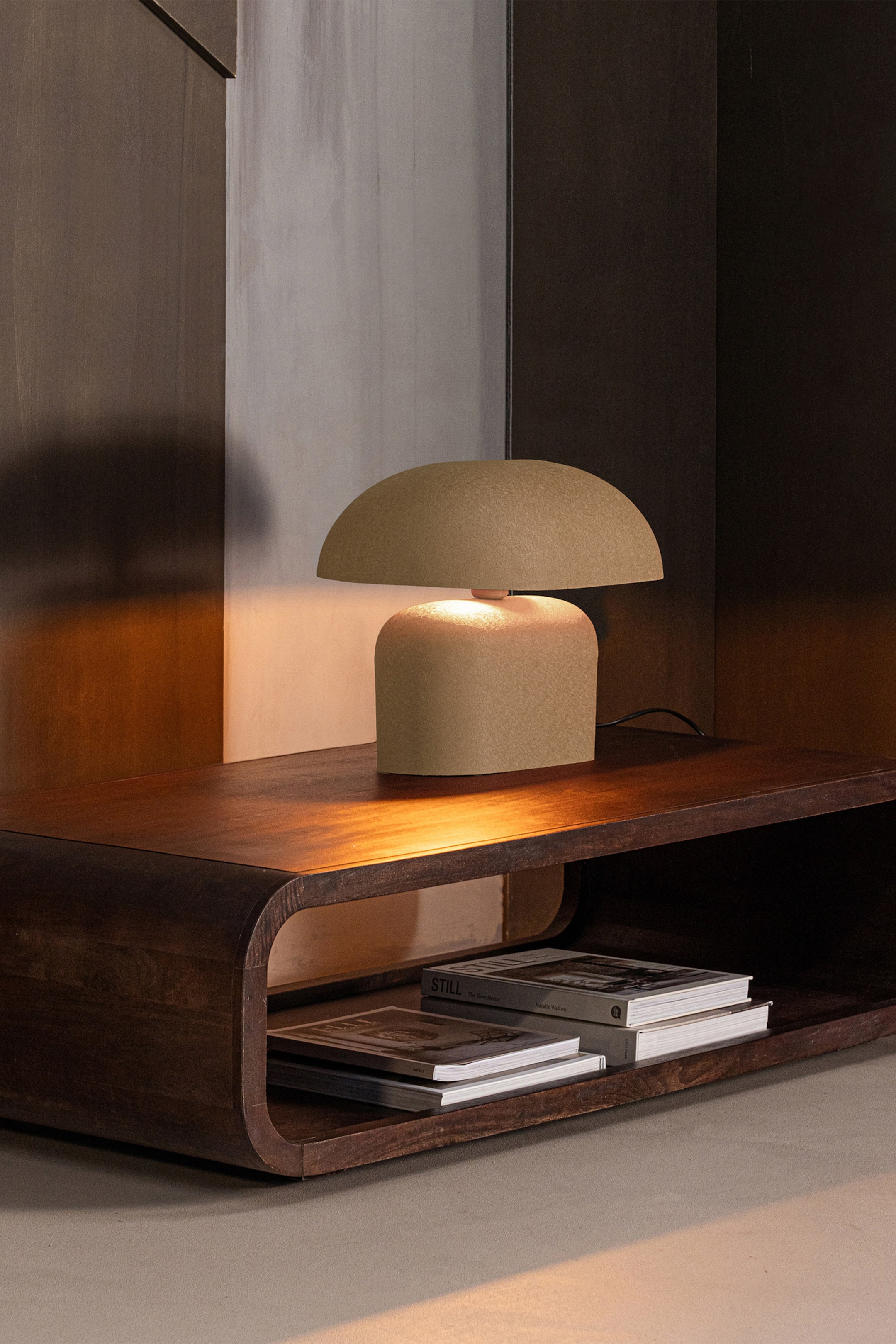 Agrandir l'image: La lampe de table texturée brun clair est positionnée à un léger angle frontal droit, avec une base rectangulaire aux coins arrondis et un abat-jour hémisphérique assorti posé au-dessus. Elle diffuse une lueur chaleureuse, avec un cordon d'alimentation noir visible à l'arrière.