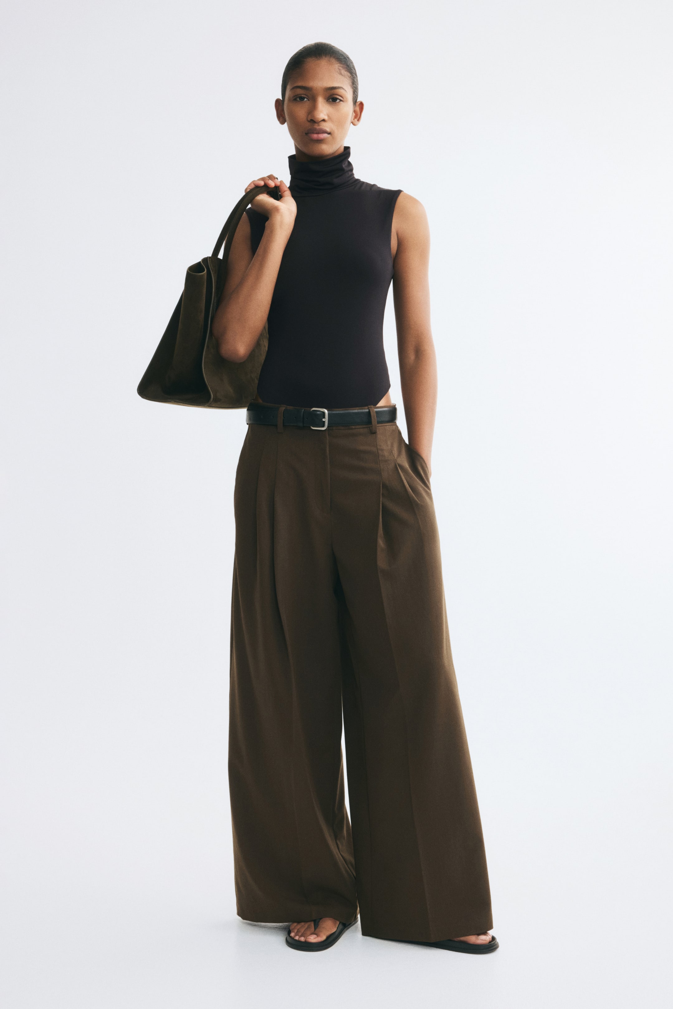 View larger image: The model is wearing brown wide-leg pleated trousers secured at the waist with a black leather belt. The trousers are paired with a sleeveless black turtleneck bodysuit, and a dark olive-green suede tote bag is carried over the shoulder.