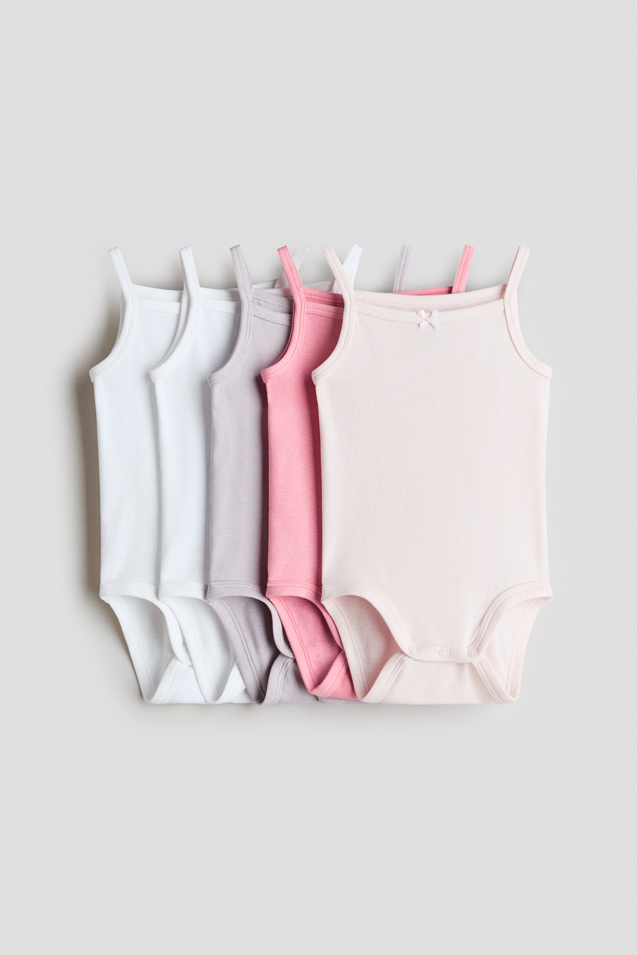View larger image: A stack of five sleeveless bodysuits is neatly arranged, each displaying its front. These are made from soft cotton jersey, with delicate shoulder straps, scoop necklines, and press-studs at the crotch for convenience. The bodysuits come in shades of white, light grey, pink, and light pink. Notably, the light pink bodysuit is adorned with a small bow at the neckline, adding a touch of elegance.