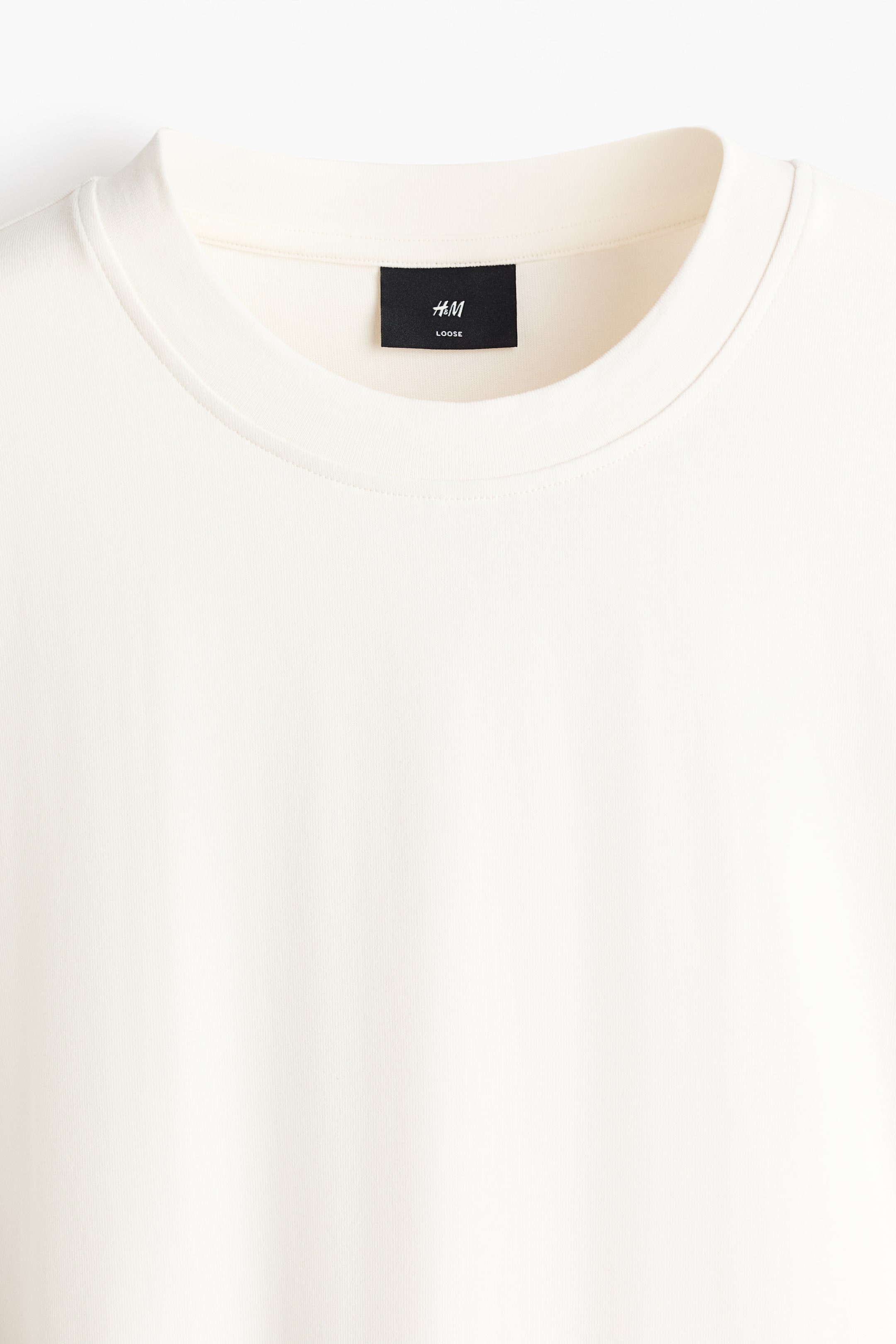 Größeres Bild anzeigen: Ein cremefarbenes Kleidungsstück wird von vorne gezeigt und verfügt über einen Rundhalsausschnitt mit einer gerippten Einfassung. Ein schwarzes H&M-Etikett mit der Aufschrift „LOOSE“ ist im Nacken innen sichtbar, und das Kleidungsstück weist eine glatte Stoffstruktur auf.