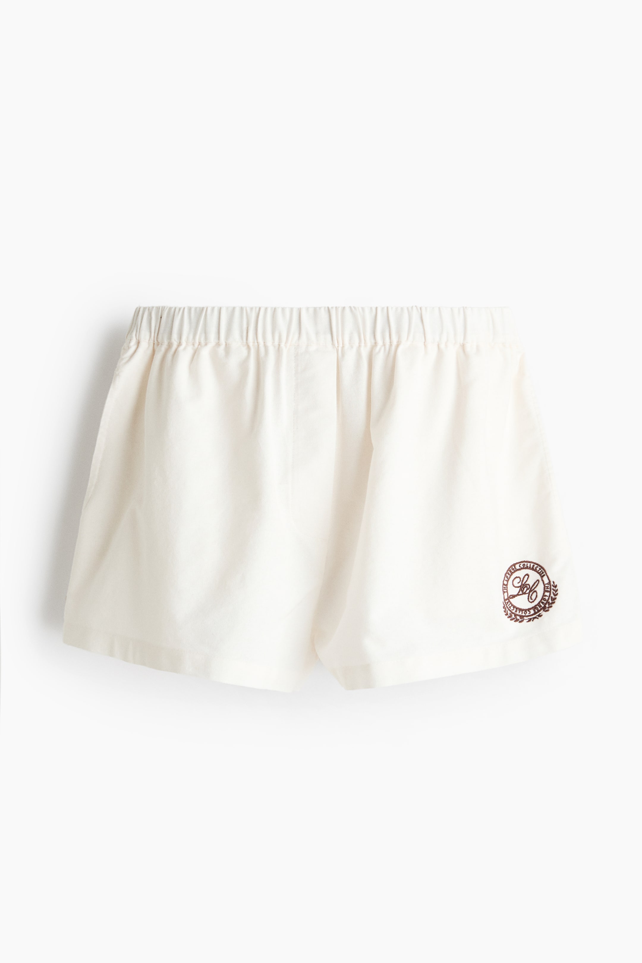 Grotere afbeelding bekijken: De off-white shorts hebben een elastische, geplooide tailleband en een donkerbruin rond embleem met tekst en een kransontwerp op de rechteronderpijp, met een ontspannen, rechte pijp snit.