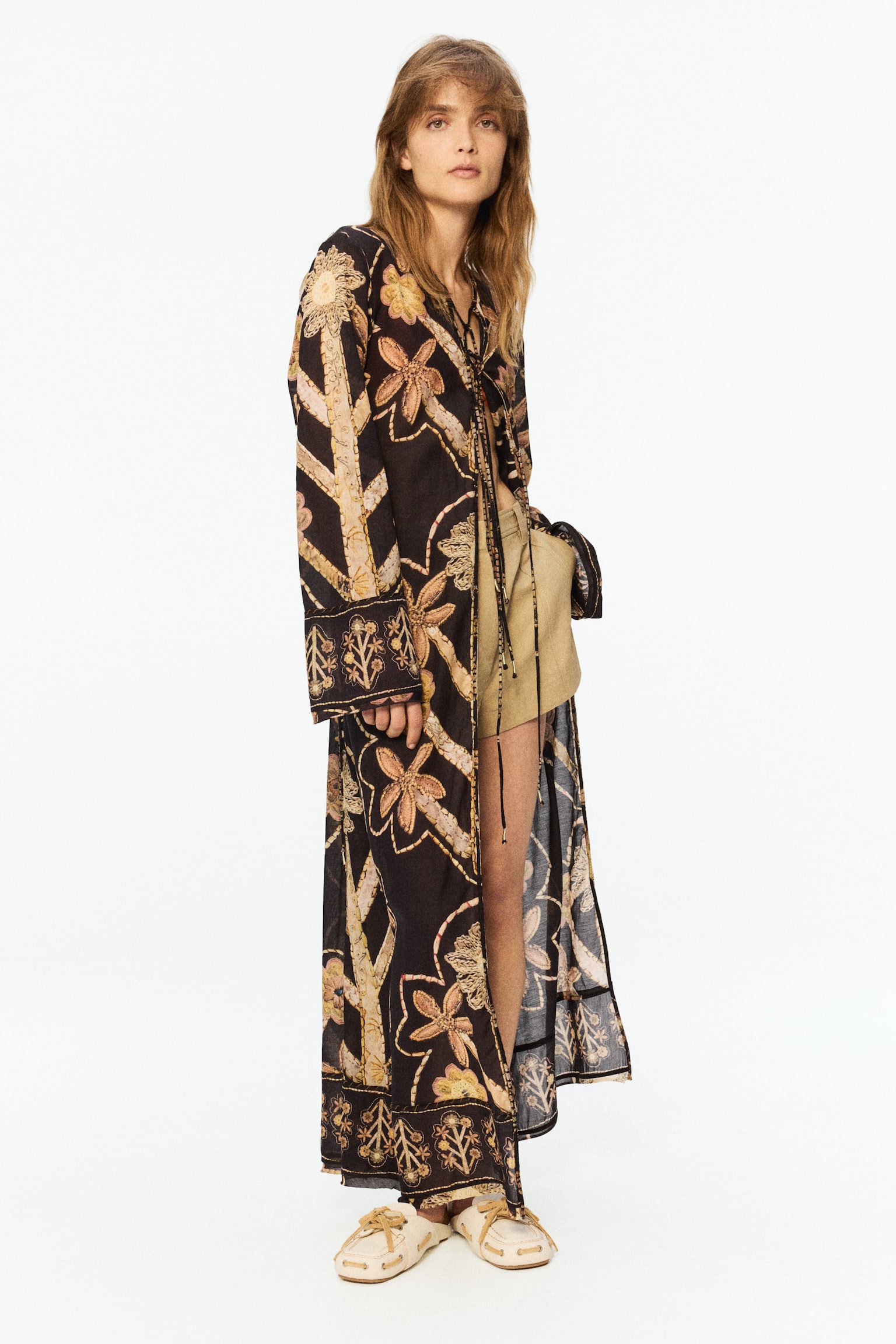 A floor-length, loose-fit kaftan dress, featuring a semi-sheer floral print on a dark background with patterned trims, is worn by the model. It has a round neckline, long wide sleeves with patterned cuffs, an open front with ties that have shimmery ends, and ties at the back cinching the waist, revealing light-coloured shorts. - 1