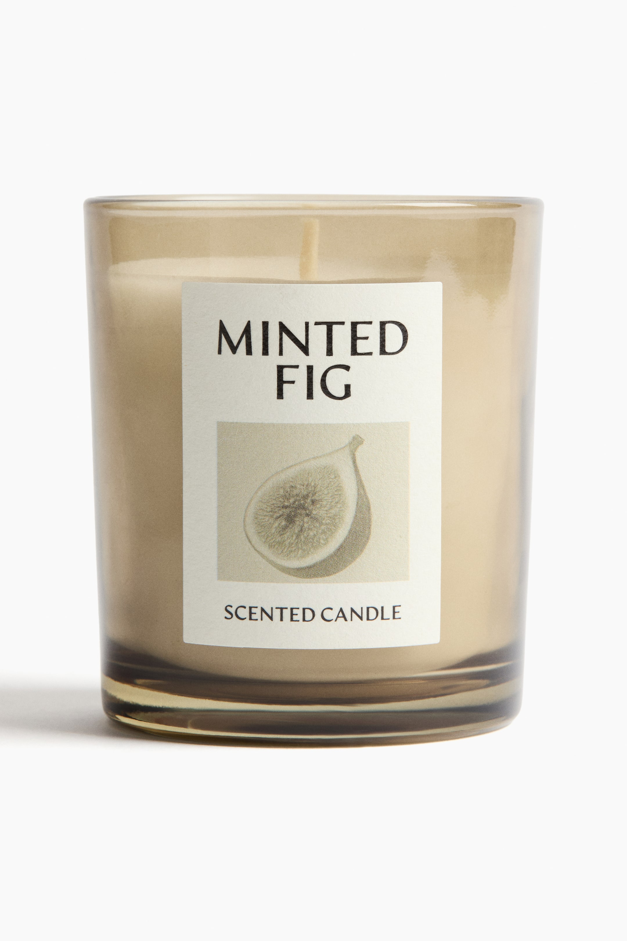 View larger image: A scented candle is presented front in a translucent light brown glass holder, containing light beige wax and a single wick. A white rectangular label is adhering to the front, displaying "MINTED FIG" and "SCENTED CANDLE" with an illustration of a halved fig.