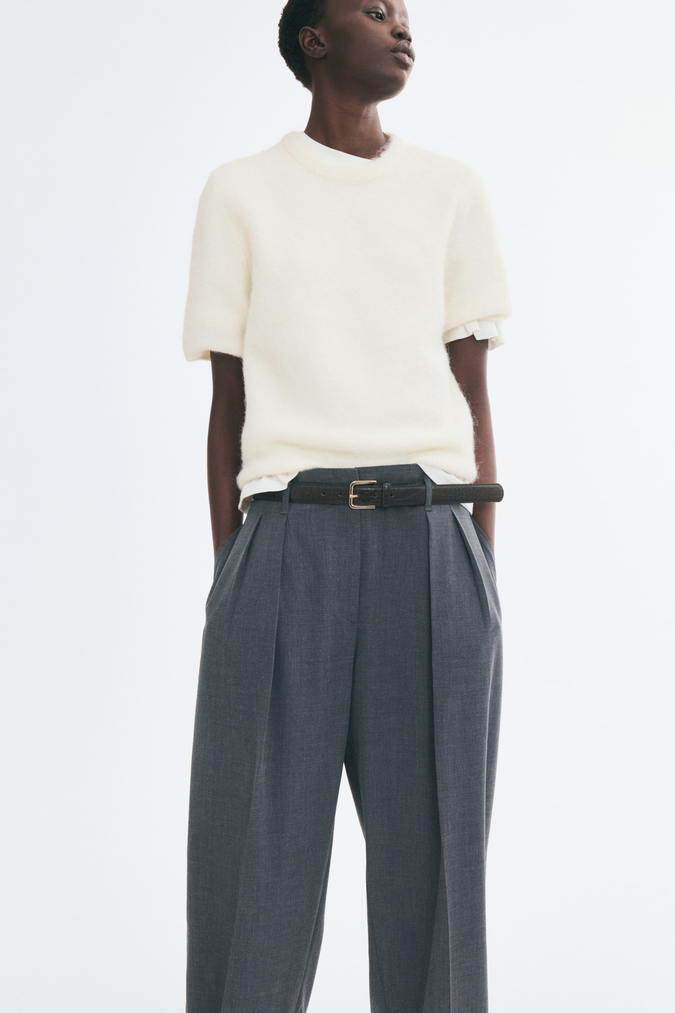 View larger image: A cream-coloured, short-sleeve sweater in a soft knit is being worn by the model over a white collared shirt. The sweater, featuring a round, rib-trimmed neckline, is tucked into grey, pleated trousers styled with a dark belt.